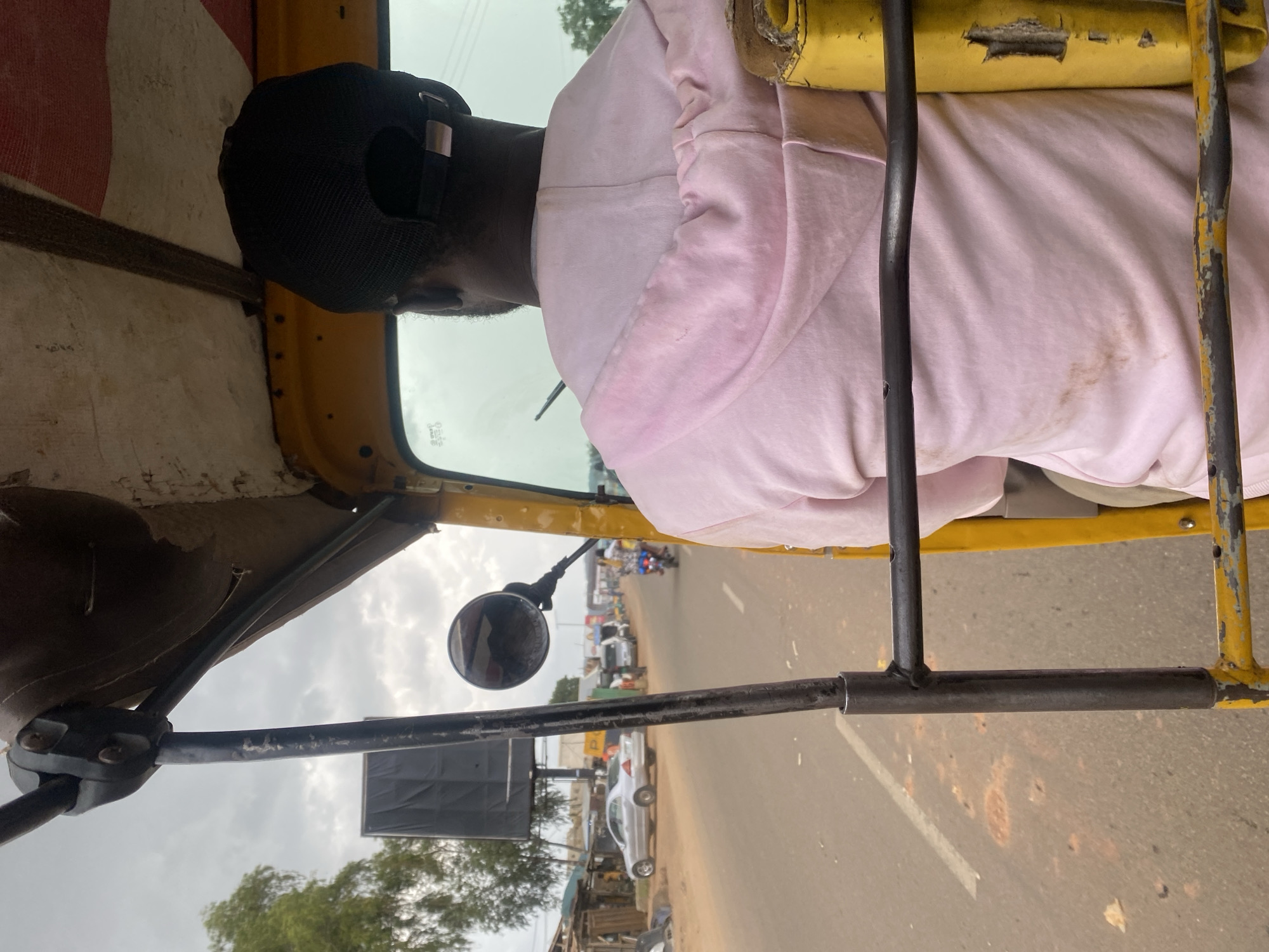 The image shows the interior view of a yellow auto-rickshaw, with the driver in the foreground. The driver, wearing a black baseball cap and a pink hooded sweatshirt, is facing away from the camera and steering the vehicle. The auto-rickshaw is moving along a paved road in Rafin Par, Nigeria, indicated by the road conditions and the style of vehicles and buildings visible. The road is lined with various establishments, including what appear to be shops or market stalls, and other vehicles, including cars and motorcycles, are present. The sky is overcast. The auto-rickshaw has a protective metal frame visible throughout the frame.