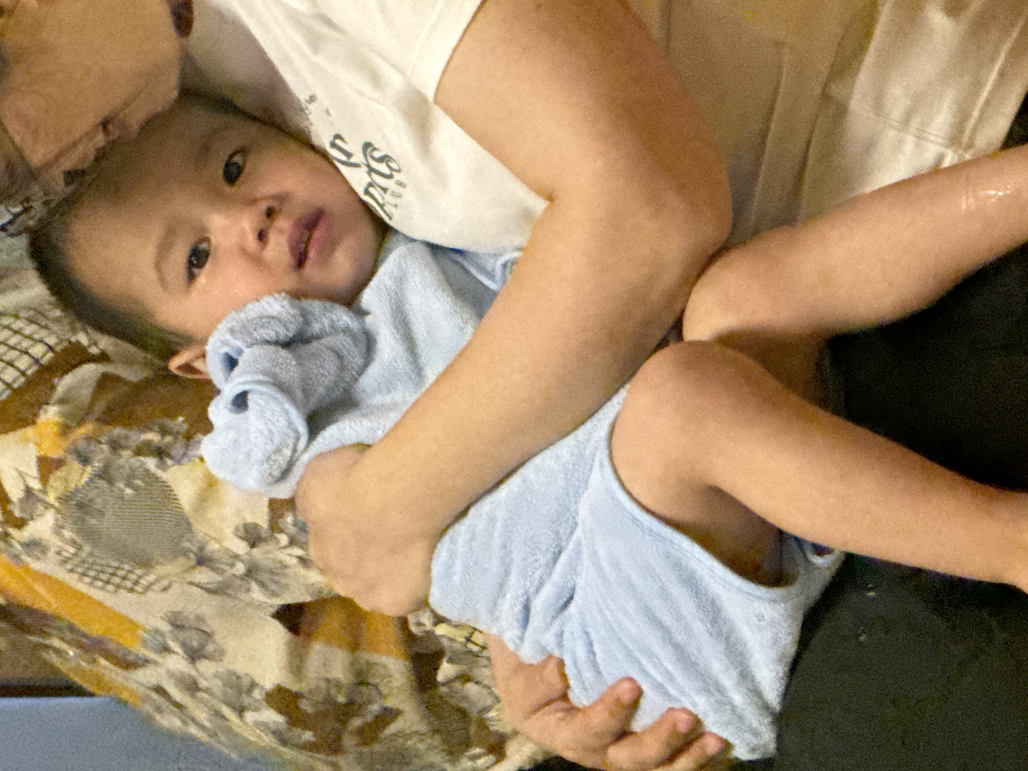 This is a close-up, eye-level, indoor photograph of a baby being held by an adult. The baby, who appears to be a toddler, is wrapped in a light blue towel and is resting on a patterned blanket with orange and brown floral designs. The baby's head is turned to the side, looking towards the viewer with dark eyes and pursed lips. The adult's arm is visible, gently holding the baby. The adult is wearing a white t-shirt with some text visible on it. The text on the t-shirt appears to be in a stylized font, possibly with Chinese characters. The lighting is soft, suggesting an indoor setting, likely in a home environment. The overall mood is one of tenderness and comfort, as the adult holds the baby close. The scene is likely taking place in Guandu, China, as indicated by the location context. There are no strong cues regarding the time of day or weather.