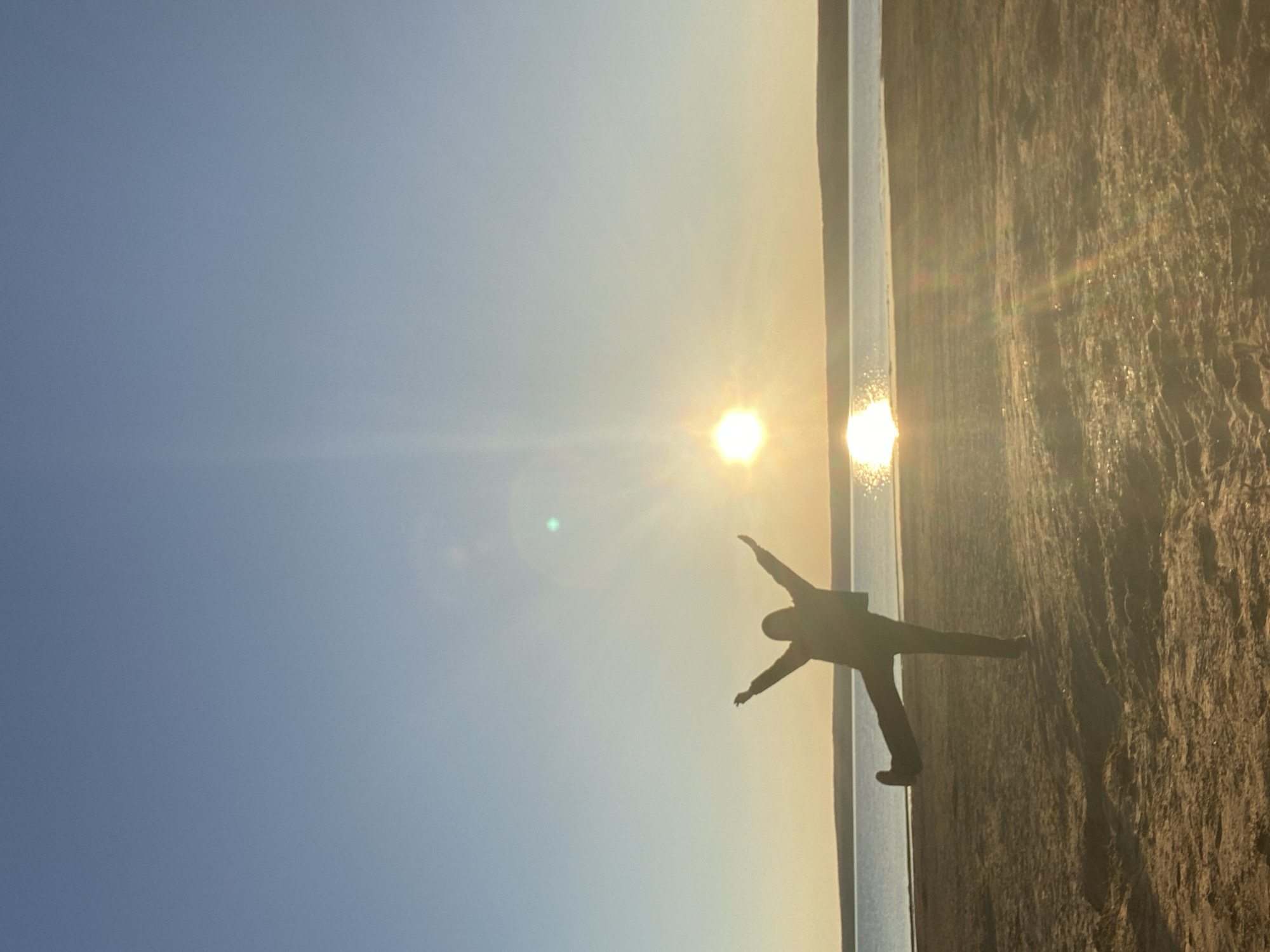 The image shows a silhouette of a person standing on a textured surface, likely sand, near a body of water. The person has their arms outstretched, appearing to be in a "starfish" pose. The sun is setting or rising over the water, creating a bright glare and long reflections. The sky is a gradient of blue to yellow, with the brighter yellow near the horizon. The location is Chaikovsky, Russia.