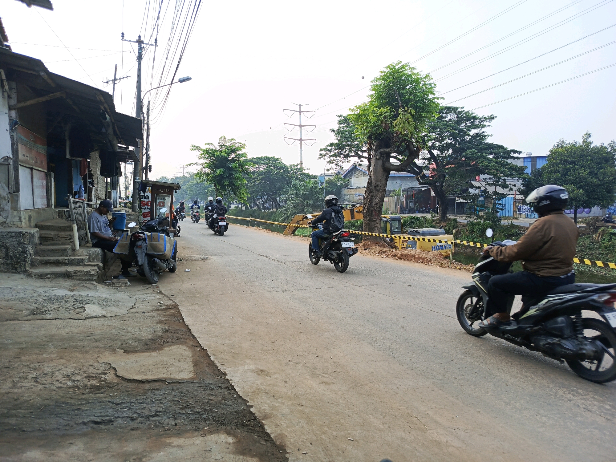 A street scene in Lebak Wangi, Indonesia, features several motorcycles and their riders on a paved road. On the left, a man in a gray t-shirt and a dark cap sits on a step next to a motorcycle with two large bags attached to the back. Adjacent to him is a weathered building with a dark, corrugated roof and a white-washed lower section. A street lamp and utility poles with numerous wires stretch across the sky. In the middle ground, a motorcycle with a rider wearing a black helmet and jacket is moving away from the camera. Further down the road, more motorcycles are visible. The right side of the road is bordered by a yellow and black striped barrier, indicating construction or road work. Behind the barrier, construction equipment, including a yellow excavator, is present. Trees with lush green foliage line the road, and in the distance, buildings and power lines are visible under a bright, hazy sky. Another motorcycle rider, wearing a brown jacket and a black helmet, is in the foreground on the right, also moving away from the camera.