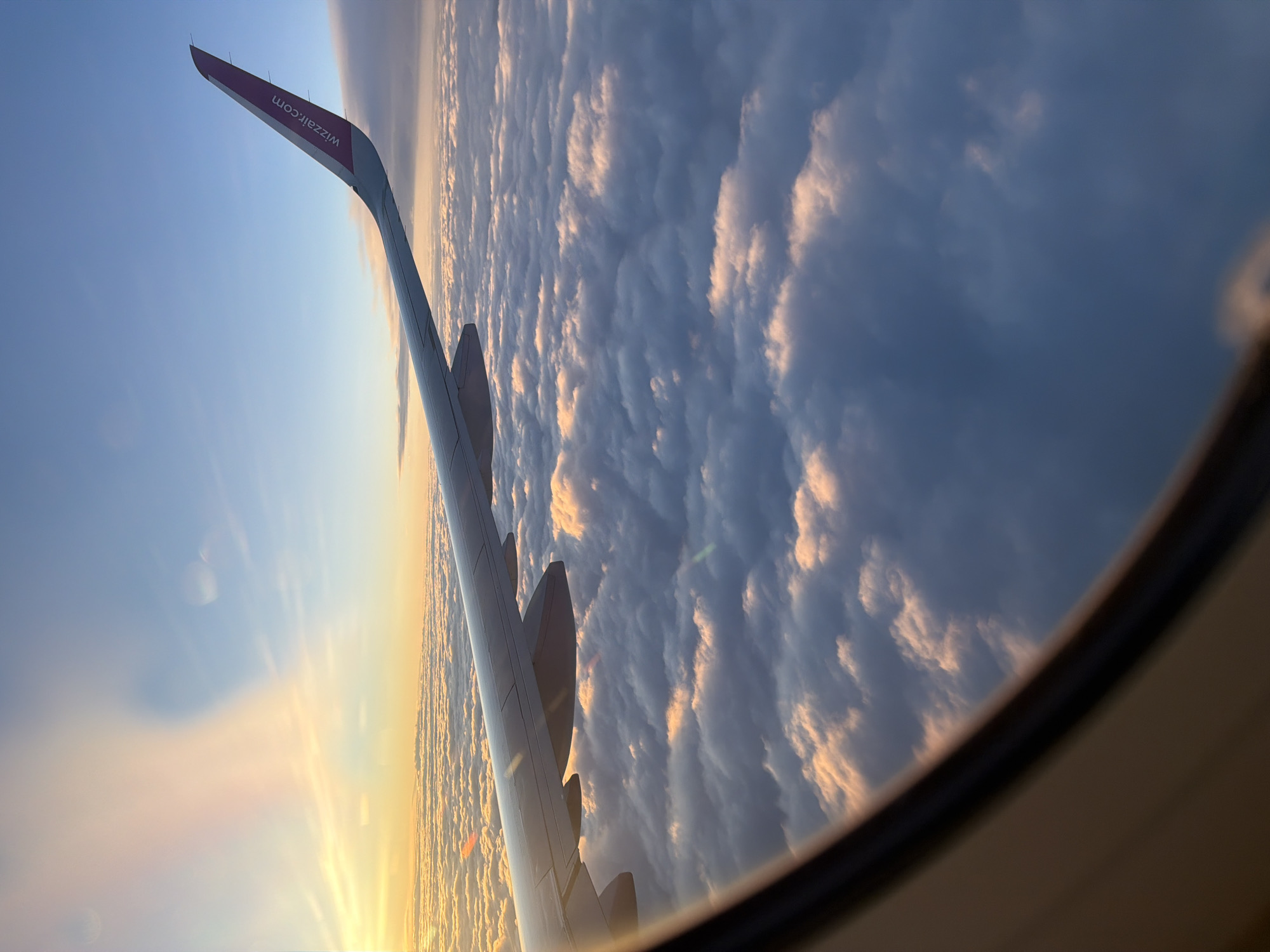 The image depicts an aerial view from an airplane window, looking out over a vast expanse of clouds. The wing of the aircraft, bearing the "wizzair.com" logo, extends into the frame from the right side. The sky is a gradient of blue, transitioning to warmer tones of orange and pink near the horizon, suggesting either sunrise or sunset. The clouds below are illuminated by this light, creating a textured and ethereal landscape. The foreground shows the interior frame of the airplane window. There is no identifiable location in Czerwony Kościół, Poland, as the image is taken from high altitude, showing only clouds and sky.