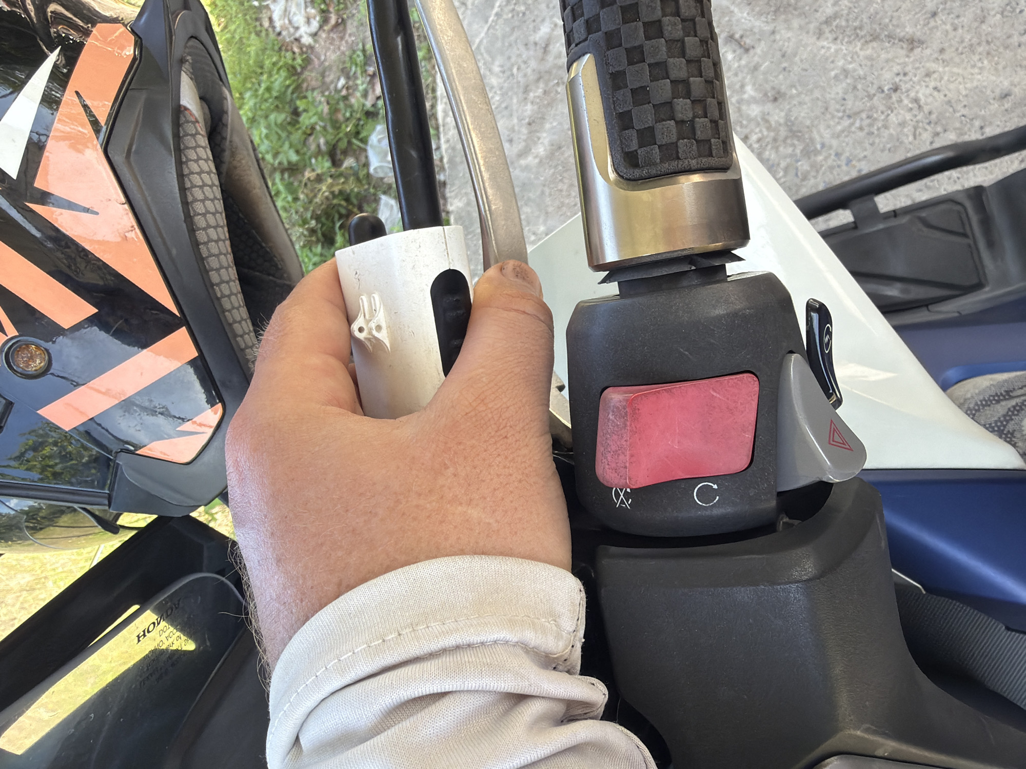 A close-up, first-person perspective shows a hand operating a motorcycle's handlebar controls. The hand, wearing a light-colored sleeve, is positioned to interact with a white toggle switch located on the left handlebar assembly. Below the switch is a prominent red button and a grey lever with a hazard light symbol. The motorcycle's black grip with a checkered pattern is visible on the right side. In the background, a portion of a black and orange motorcycle helmet is visible, along with out-of-focus green foliage and a concrete surface. The scene is consistent with a person preparing to ride a motorcycle in Baan Bo Phut, Thailand.