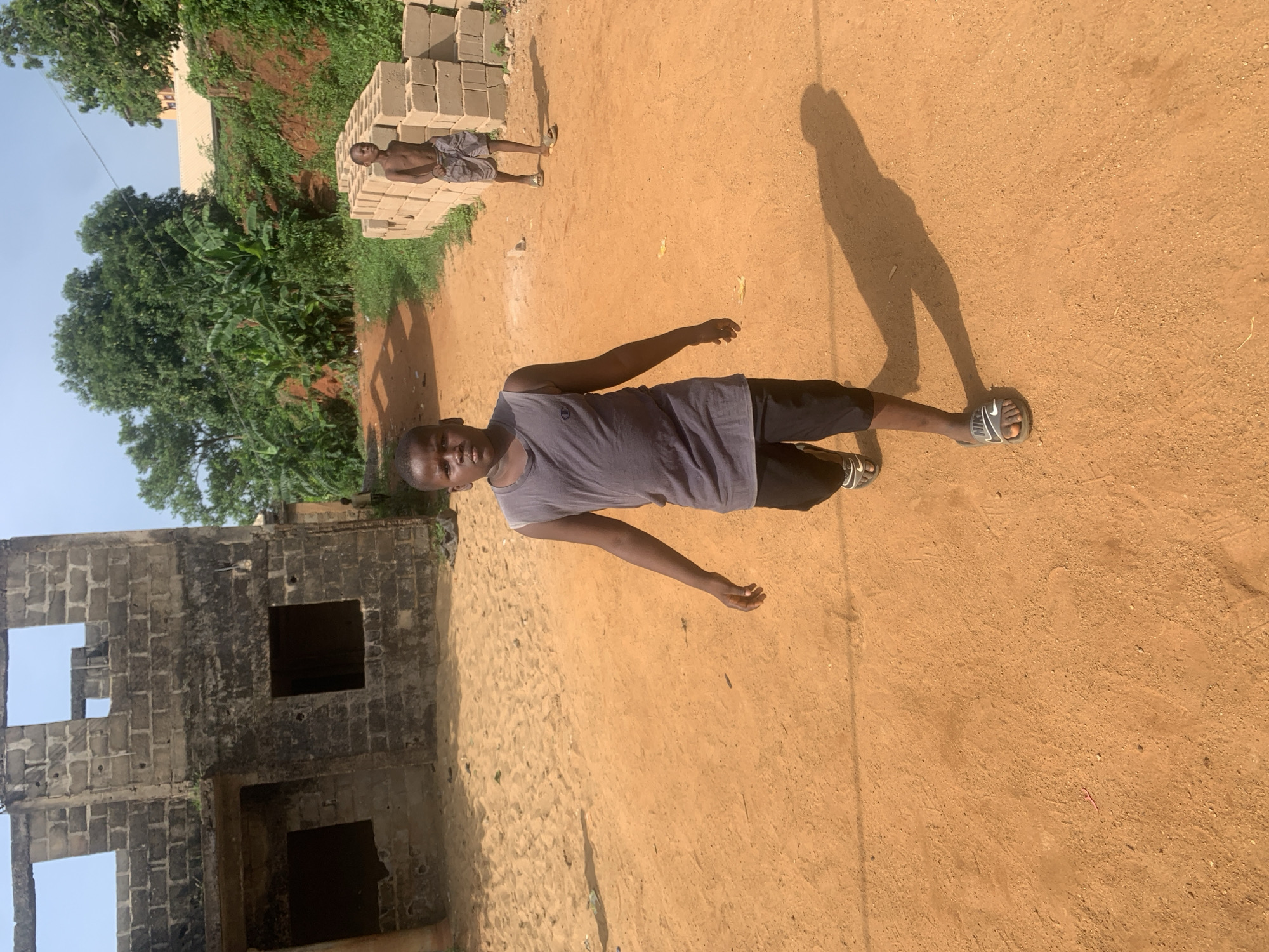 The image depicts a man standing on a dirt ground in Nsugbe, Nigeria. He is wearing a grey tank top and dark shorts, with sandals on his feet. His shadow is cast on the ground. In the background, there is a partially constructed building made of cinder blocks. To the left, a lush green area with trees can be seen. Further back, another man is standing near a stack of cinder blocks. The ground is dry and reddish-brown, typical of the soil in the region. The sky is visible in the upper left corner and is a clear blue.