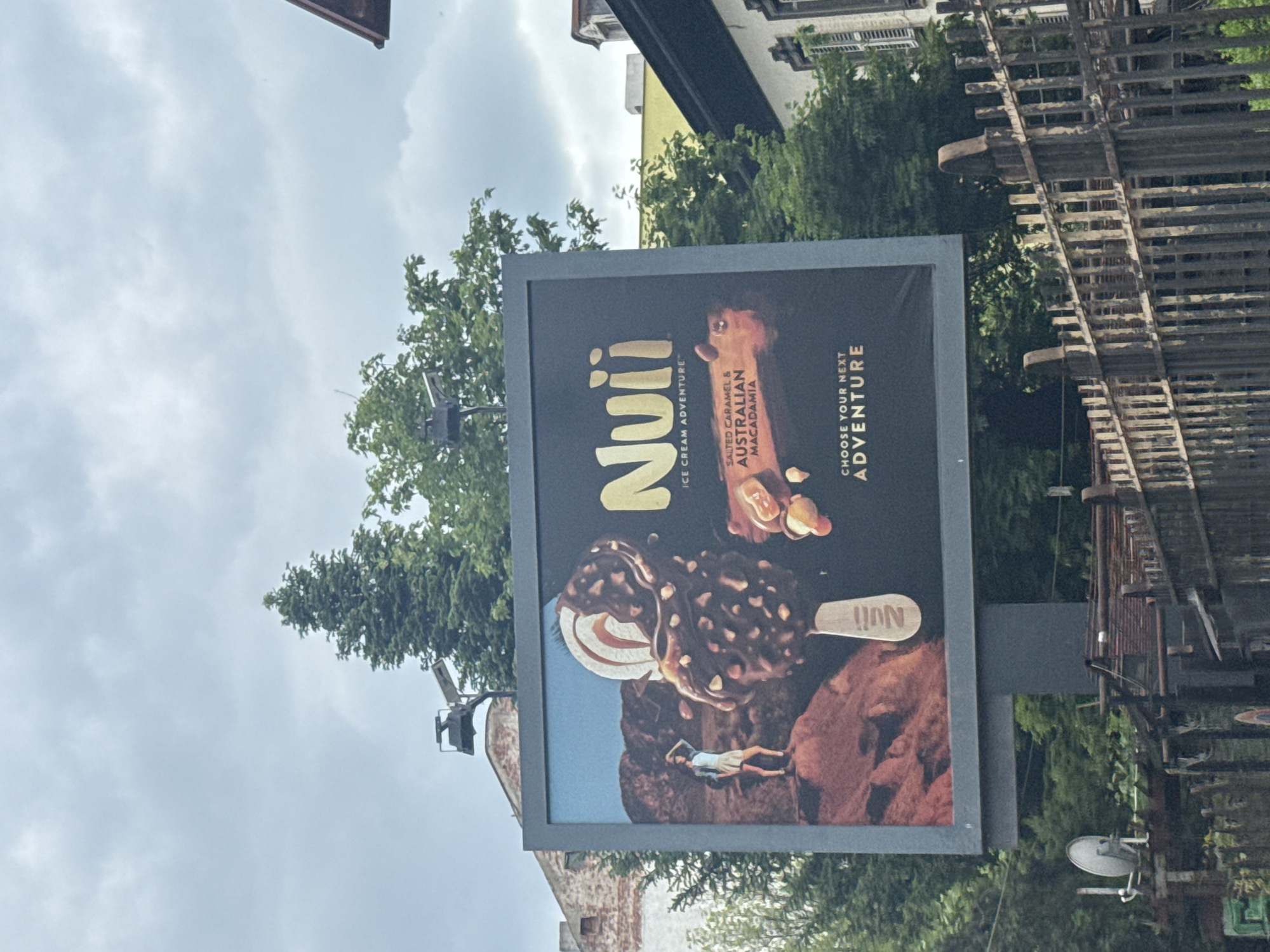 The image shows a Nuii ice cream advertisement on a billboard. The billboard displays images of Nuii ice cream bars, specifically Salted Caramel & Australian Macadamia. The lower part of the advert features a landscape background with a person lying on a rocky terrain.
The location is likely Sofia, Bulgaria. The surrounding environment includes buildings, trees, and what appears to be a fence or railing. The sky is overcast. A streetlamp is also visible near the billboard. The photograph is taken at an angle, with the billboard slightly tilted in the frame.