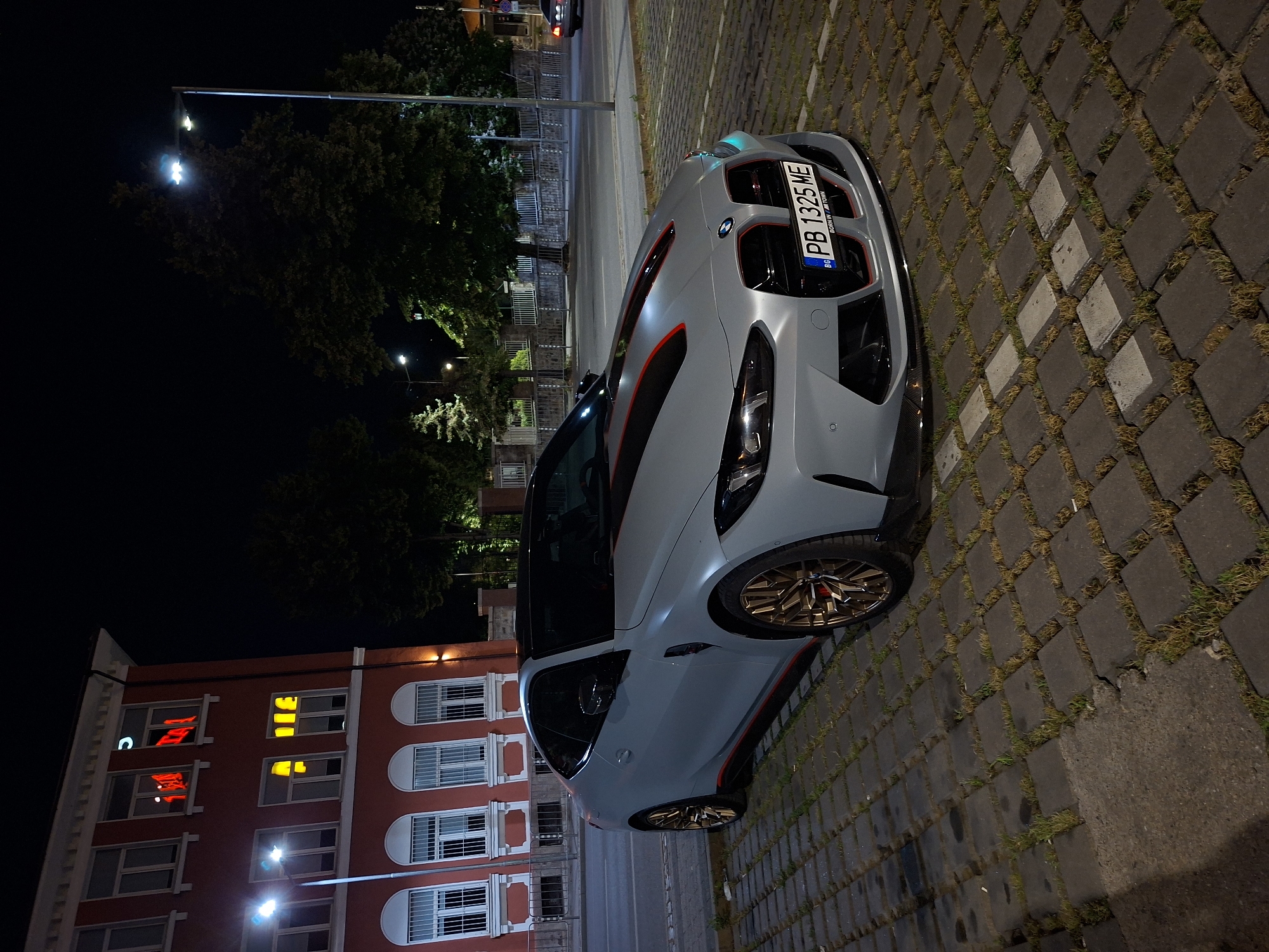 The image shows a light gray BMW M4 coupe parked on a paved lot at night in Plovdiv, Bulgaria. The car has red and black racing stripes on the hood and red accents around the grille and along the side. The license plate reads "PB 1325 ME". In the background, a red building with lit windows is visible. Trees and streetlights are also present, suggesting an urban setting. The pavement is made of square interlocking concrete blocks with grass growing between them. The overall scene is illuminated by artificial lights, creating a nighttime ambiance.