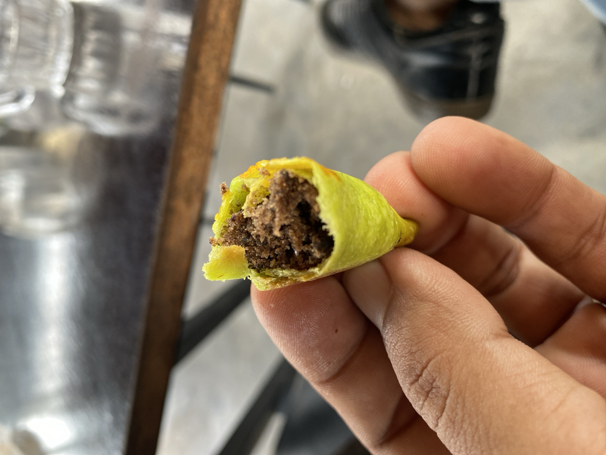 A close-up shot shows a person's thumb and forefinger holding a small, bright yellow, cone-shaped snack. The snack appears to be made of a crispy outer shell, and it is filled with a dark brown, crumbly substance. The background is blurred, but it suggests an indoor setting with a dark, wooden table and some metallic elements. Further in the background, a person's shoe is visible, indicating the presence of another individual. The overall lighting is natural and diffused, highlighting the texture and details of the snack and the hand. The scene is set in Lhokseumawe, Indonesia.