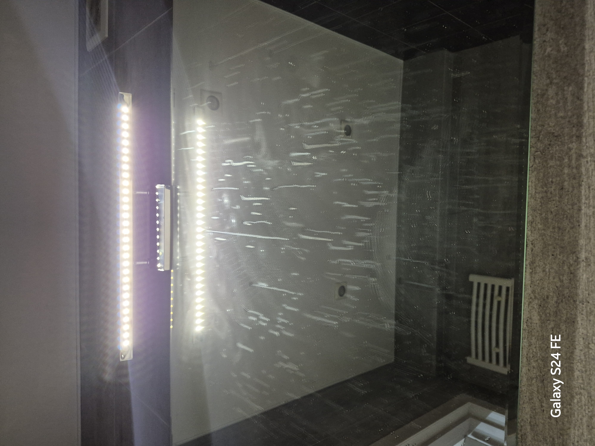 The image shows an overhead view of a shower enclosure, likely in a residential bathroom. A bright LED light fixture is positioned above the enclosure. The glass walls of the shower are streaked with water marks and appear slightly fogged. Inside the enclosure, the bottom of a shower head or faucet is visible, along with a slatted drain cover on the floor. The background consists of dark tiled walls and a ceiling. The bottom of the image contains a watermark that reads "Galaxy S24 FE". There are no people or active interactions depicted in the scene. The setting does not provide any specific clues to indicate it is located in Chișinău, Moldova.