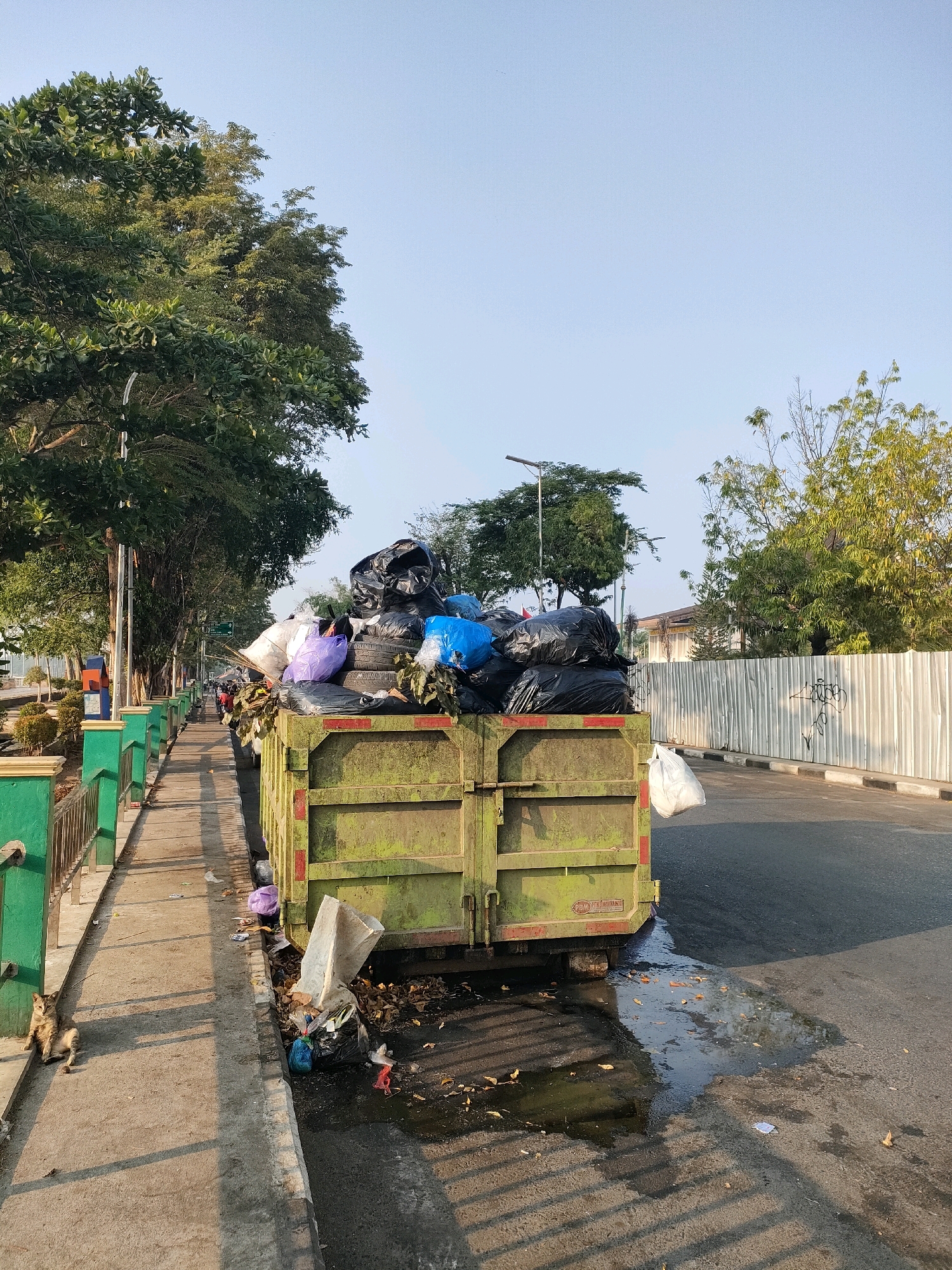 A full-frame, eye-level outdoor medium shot captures a garbage truck overflowing with black trash bags, tires, and miscellaneous refuse. The truck is positioned on the right side of the frame, adjacent to a sidewalk. The truck's body is a faded olive green, showing signs of wear and tear. Scattered trash and leaves litter the ground around the truck.
To the left of the truck is a concrete sidewalk with a green metal railing. A small, tabby cat is seated on the sidewalk, facing left, in the lower left quadrant of the image. Beyond the sidewalk, a row of trees lines a park-like area with decorative fencing.
In the background, a white metal fence runs along the right side of the road. The sky above is clear and pale blue, suggesting an early morning or late afternoon setting. The scene appears to be located on a street in Banjarmasin, Indonesia, given the presence of local flora and urban elements. The overall impression is one of urban waste management in a tropical environment.