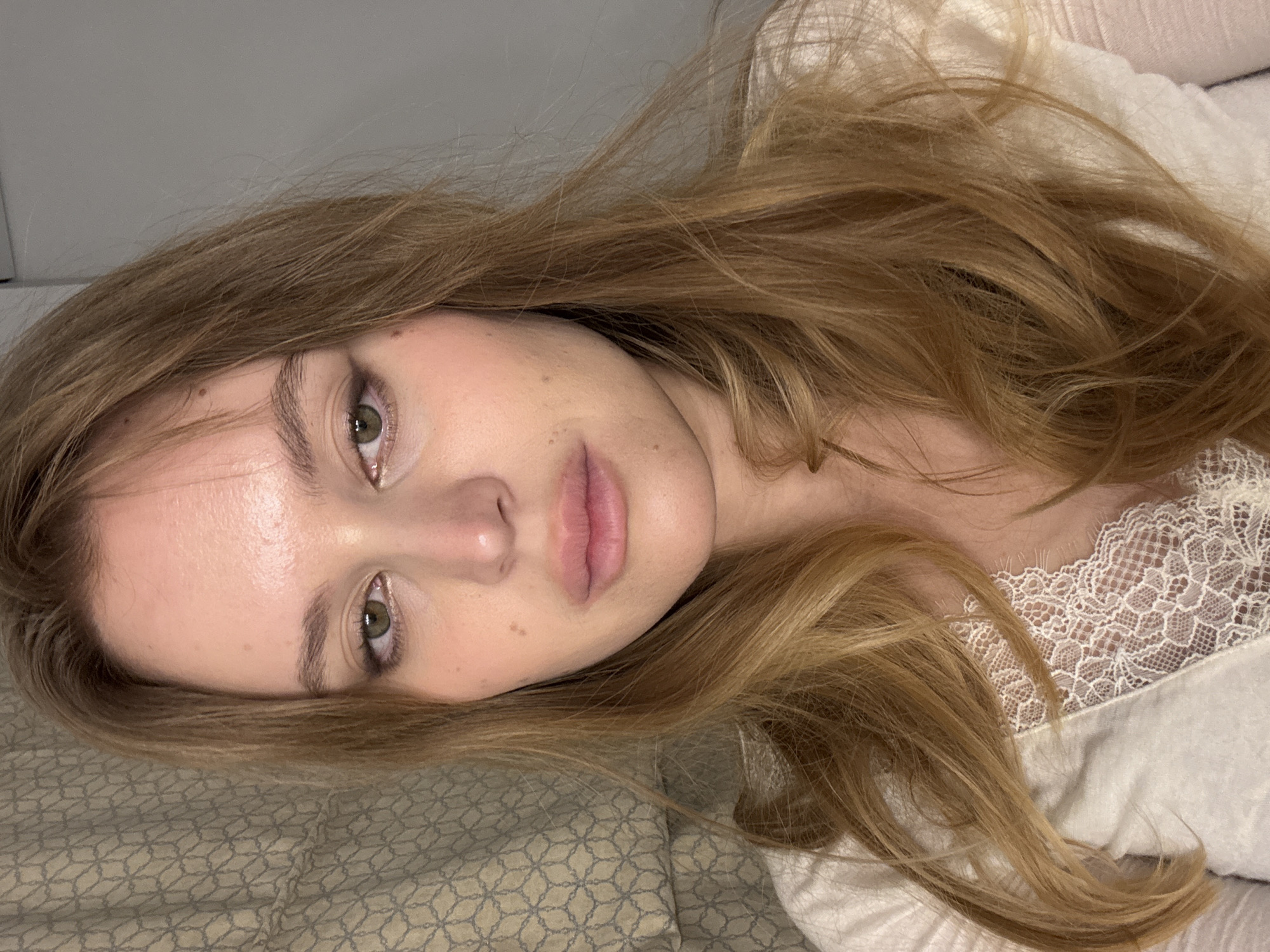 A young woman with long, light brown hair is lying down. Her head rests on a patterned pillow or bedsheet, and she looks directly at the camera. She has fair skin, green eyes, and full lips. She is wearing a white top with a lace trim. The background appears to be a plain wall. The location is not discernible from the image, so a location in Hrodna, Belarus, cannot be determined.