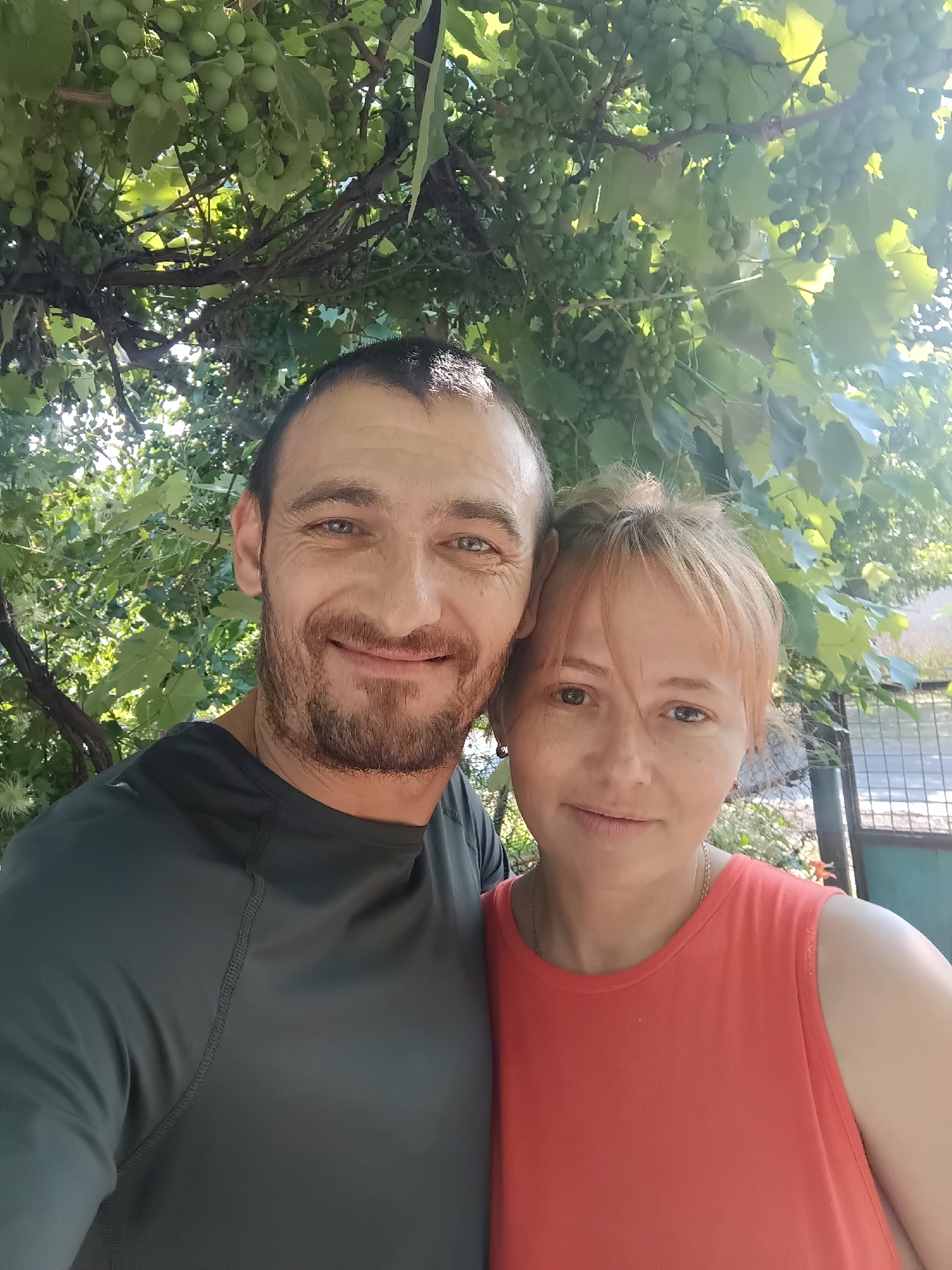 A selfie taken outdoors shows a man and a woman smiling and looking at the camera. They are positioned in the foreground, with lush green grapevines and leaves filling the upper half of the image. The man, on the left, has short dark hair, a beard, and is wearing a dark gray long-sleeved shirt. The woman, on the right, has blonde hair styled up and is wearing a sleeveless coral-colored top. Sunlight filters through the leaves, creating a bright, dappled effect. In the background, out of focus, a chain-link fence and some vegetation are visible, suggesting an outdoor setting possibly in a yard or garden. The scene is likely in Saksahan, Ukraine, given the context of the request.