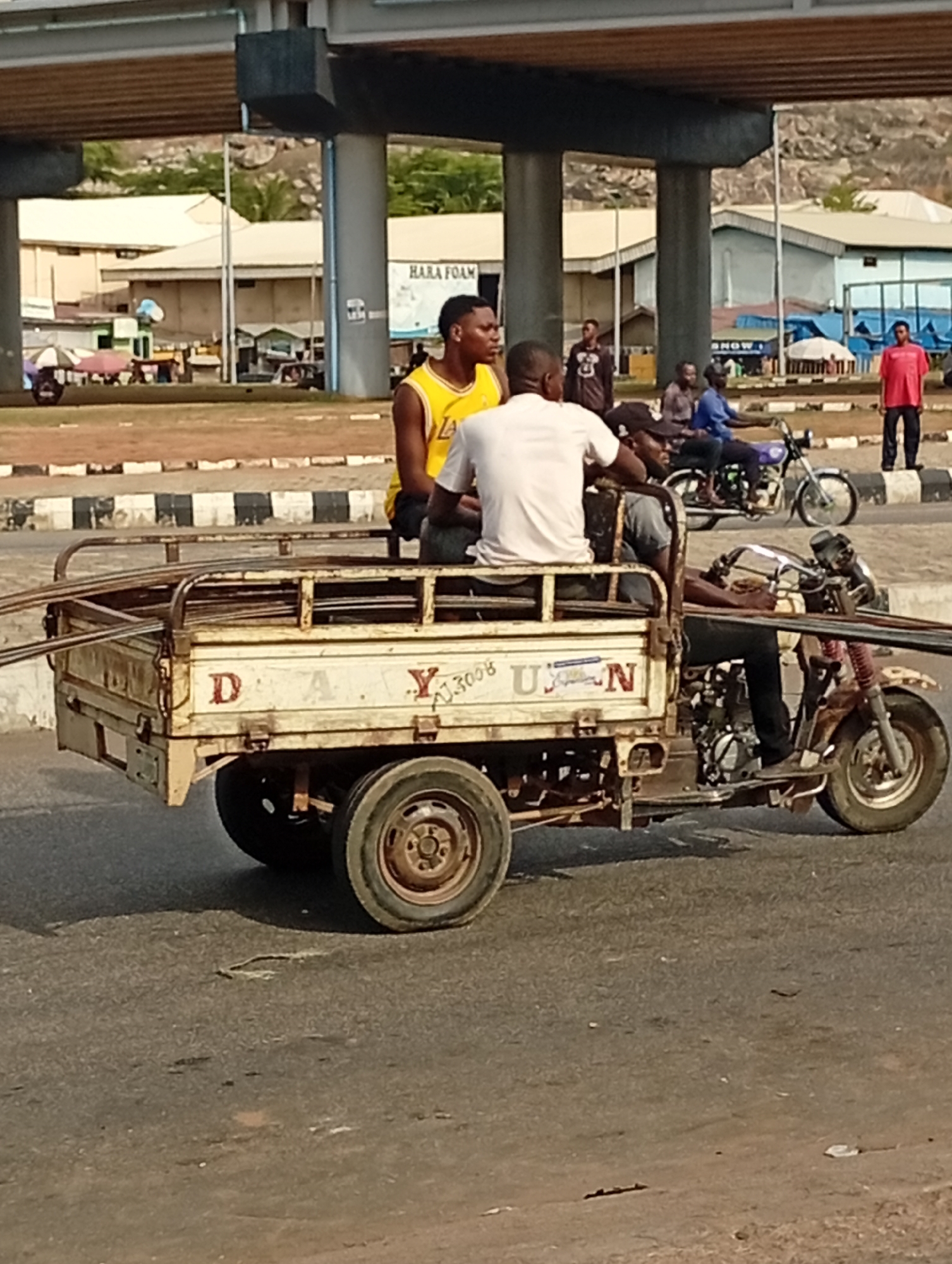 The image depicts a motorized tricycle in Lokoja, Nigeria, on a paved road, with three men riding on it. The tricycle has a cargo bed, with the letters "D-A-Y-U-N" painted on the side. The location is beneath an elevated concrete highway bridge. In the background, there are buildings, trees, and a mountain range. Other individuals can be seen standing and sitting along the roadside, as well as a motorcyclist. A sign reads “HARA FOAM”.