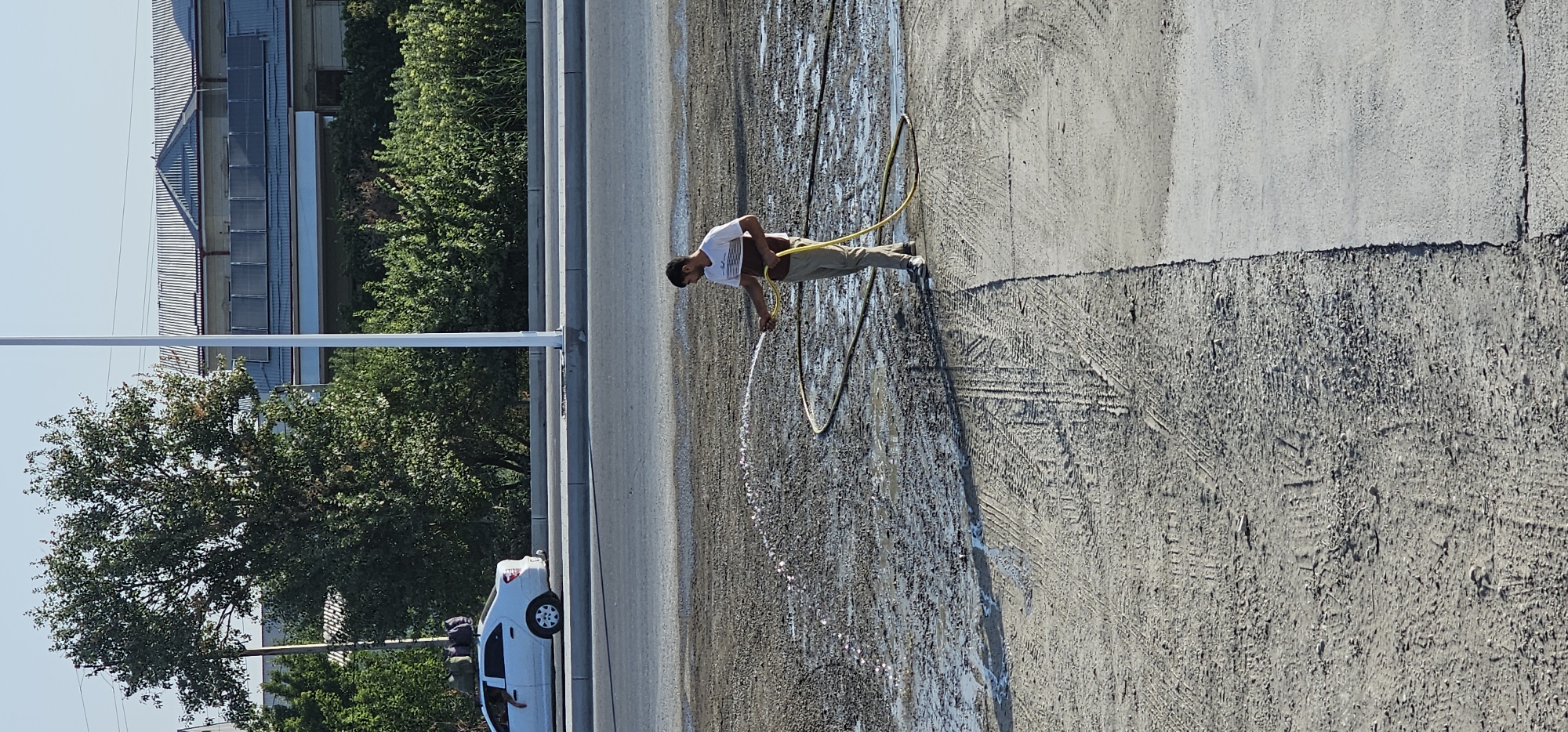 In the city of Sheyhancha, Uzbekistan, a man is seen watering a construction site with a hose. He is wearing a striped t-shirt and beige pants. The ground is muddy and appears to be a mixture of dirt and gravel, with tire tracks visible in the foreground. In the background, a white car is parked on the side of a road, with luggage strapped to its roof. Beyond the road, lush green trees and a building with solar panels on its roof are visible under a clear blue sky. The scene suggests ongoing construction or maintenance work.