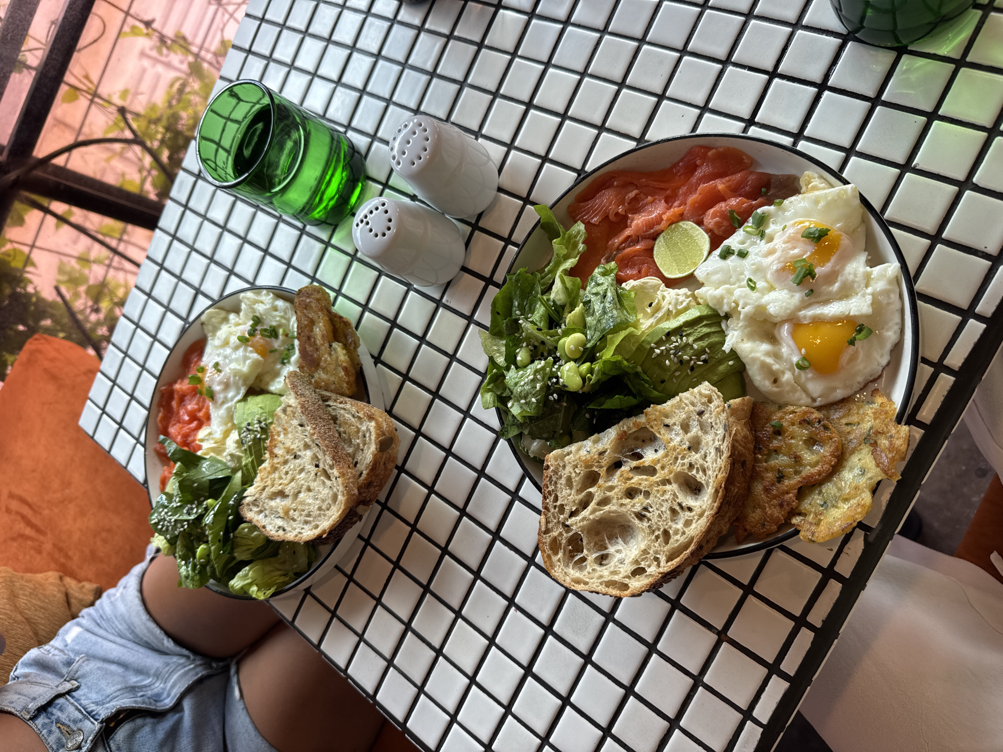The image captures an overhead shot of two bowls of food placed on a table with a white tile surface. Each bowl contains a salad with salmon, avocado, greens, fried eggs, and slices of bread. Additionally, one bowl includes fried latkes. Salt and pepper shakers and a green glass are positioned on the table next to the bowls. A person wearing denim shorts is seated beside the table with one of the bowls of food in front of them. The location is North Kuta, Indonesia.