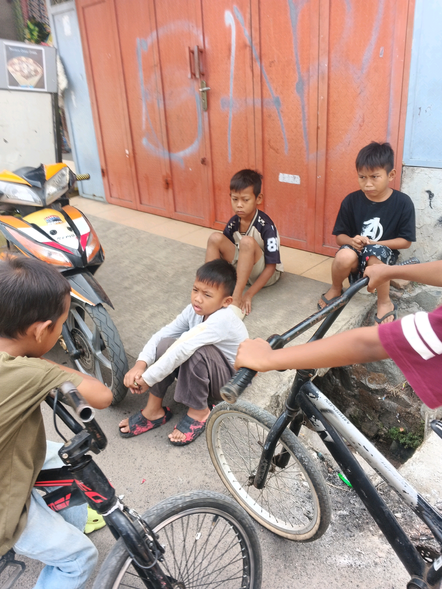 In Kadu, Indonesia, four young boys are gathered on a paved surface in front of a large, orange, corrugated metal garage door. The door is graffiti-marked with white spray paint. Two boys sit on the concrete steps leading up to the door, while two others are seated on the ground. A yellow and black motorcycle is parked to the left of the frame, and two bicycles are in the foreground. One boy on the right wears a black t-shirt with a white logo and patterned shorts, while the boy next to him wears a grey long-sleeved shirt and dark pants. The two boys on the steps are wearing a blue and white striped shirt with the number 81 on it, and a tan t-shirt, respectively. The boy on the far left wears a green t-shirt. The overall scene depicts a casual gathering of children with their vehicles on a street.