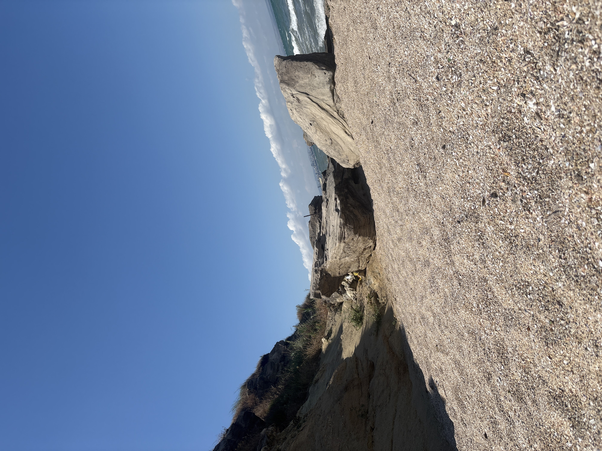 The image captures a scenic view of a rocky coastline under a clear blue sky. The foreground is dominated by a textured surface of sand and shells, creating a natural embankment. Beyond this, weathered rocks of various sizes form a rugged landscape, with some jutting out towards the sea. In the distance, the ocean meets the horizon, with waves crashing against the shore and white clouds streaking across the sky. Hints of vegetation, like dry grasses and reeds, grow on the slopes of the rocky terrain. There are two small, unidentifiable objects scattered among the rocks, appearing as yellow and white debris. The lighting suggests it is daytime, likely sunny, given the clear sky and the shadows cast. The location appears to be a coastal area in Burgas, Bulgaria, characterized by its dramatic rock formations and proximity to the sea. No people are visible in the frame, and there is no discernible text. The overall impression is one of a wild and natural coastal environment.