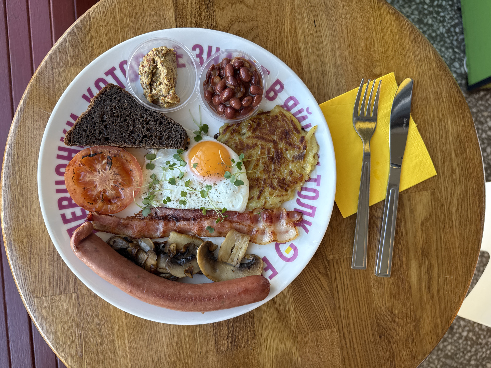 A bird's-eye view shows a breakfast plate on a round wooden table. The plate contains a variety of items: a slice of dark bread, a small container of mustard, a grilled tomato slice, microgreens, a fried egg, two strips of bacon, sliced mushrooms, a sausage, a container of beans, and a potato pancake. A fork and knife rest on a yellow napkin next to the plate. The table surface has a wood grain pattern. The photo was likely taken in Saint Petersburg, Russia, based on the Cyrillic text visible on the plate, though the details of the text are not clear enough to provide specific locational clues within the city.