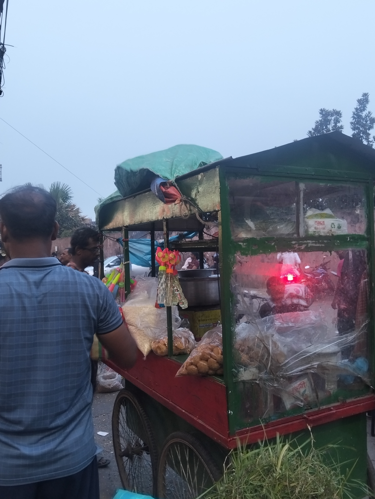 The image captures a street scene featuring a food vendor's cart, likely selling snacks like pani puri or puchka, common in South Asia. The cart is constructed from wood or metal, painted red and green, and has a green tarp covering its roof. Clear plastic sheeting encloses parts of the cart, revealing bags filled with various food items such as puffed rice and small, round, fried shells. Small decorative toys or trinkets hang from the cart's structure.
In the foreground, a man with a partially bald head and a blue checkered shirt stands with his back to the camera, appearing to interact with the vendor or wait for service. Another man with glasses is visible behind him, facing sideways. Other blurry figures are present in the background, suggesting a moderately busy outdoor setting.
Through the clear panel of the cart, a motorbike with a bright red tail light is visible. The sky is overcast and hazy, indicating either early morning, late afternoon, or evening, contributing to the subdued lighting. Green foliage and trees are seen in the background and at the base of the cart.
Visible text on packaging inside the cart includes what appears to be Bengali script on a white package (possibly "সুবর্ণা" or a similar brand name), accompanied by what resemble recycling symbols. Another yellow box label partially reads "RUBBER SOLUTION" and "Cobra Weight." The scene depicts a typical street food experience.