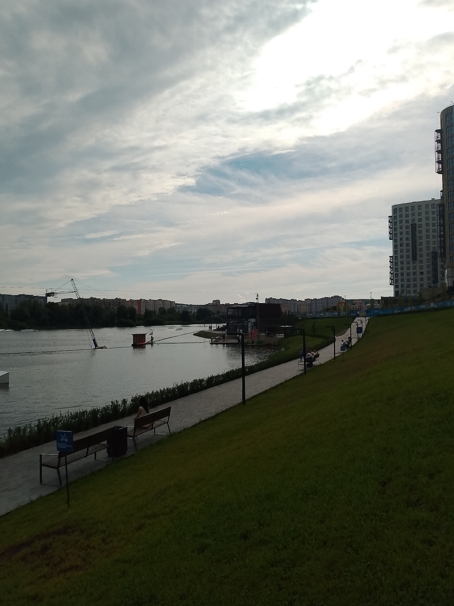 The image depicts a watersports facility on a lake in Penza, Russia, with modern buildings and a park area in the foreground. A cable system for wakeboarding is visible across the water, with participants engaged in activities. Along the water's edge, a paved walkway is lined with benches and greenery. A grassy slope with a walking path curves away from the viewer into the distance, where more residential buildings are seen. The sky is cloudy.