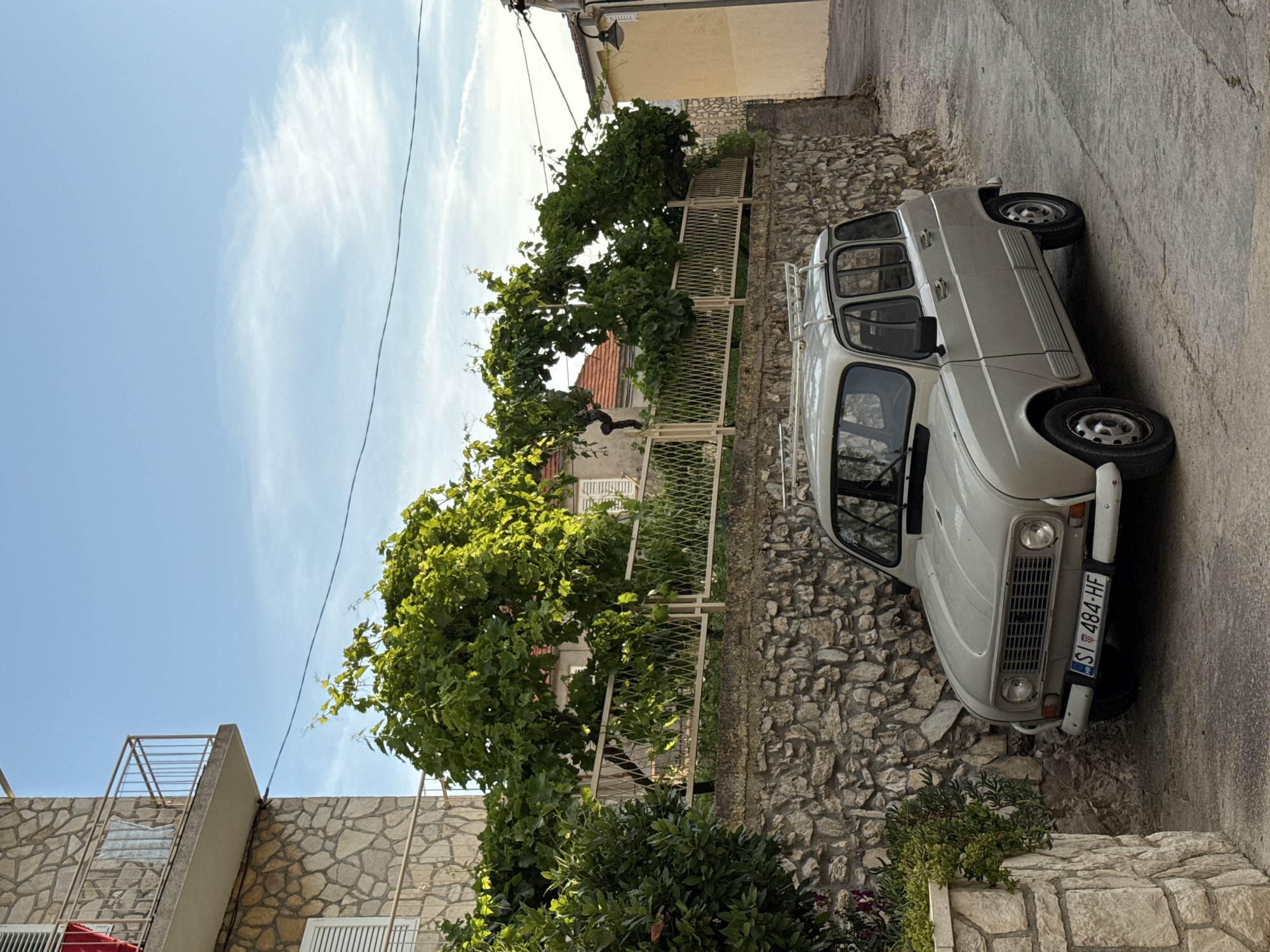 A light gray, vintage Renault 4 car is parked on a narrow, paved street in Primošten, Croatia. The car is positioned at an angle, facing towards the viewer's left. The street slopes gently downwards from right to left. To the left of the car, a rough stone wall runs along the property line, topped with a metal fence. Lush green foliage, appearing to be grapevines and other bushes, grows behind the fence and cascades over the wall in places. A multi-story building with stone cladding and a balcony is visible on the far left. The sky above is a clear blue with some wispy white clouds. The overall atmosphere is serene, suggesting a quiet residential area.