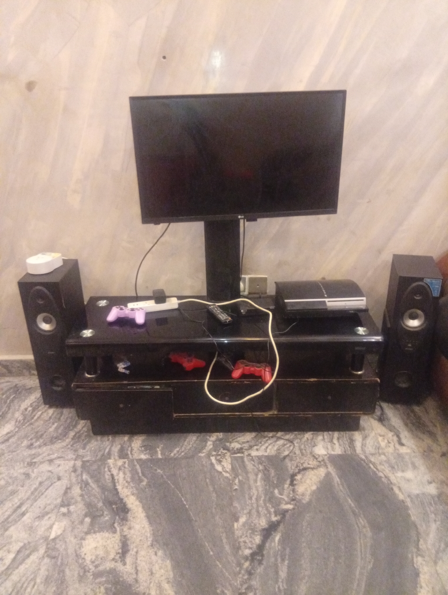 The image depicts a room in Aviation Quarters, Nigeria, featuring a flat-screen television mounted on a stand atop a black glass TV stand. A Playstation 3 console sits on the right side of the stand. A purple Playstation controller and an electrical power strip are positioned on the left side of the stand. A white object is seen on top of the left speaker, and a red Playstation controller is visible on the shelf below. Speakers flank either side of the TV stand. The floor is tiled with a marble-like pattern, and the wall behind the setup is textured with a neutral color. There is no indication of any person present in the image. The scene showcases a simple entertainment setup.
