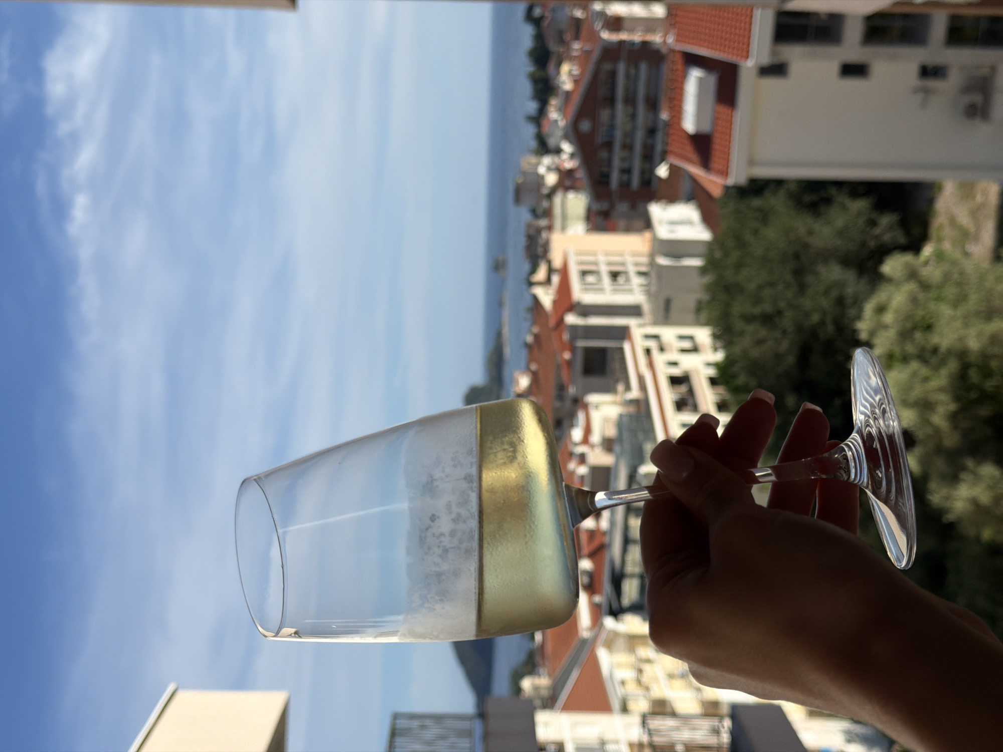 A hand holds a wine glass filled with a pale yellow liquid against a backdrop of a coastal city. The glass, tilted, shows a frothy top layer and a clear liquid below, catching the sunlight. In the background, a cluster of buildings with red tile roofs stretches towards the sea. Beyond the city, a body of water meets the horizon under a bright blue sky dotted with wispy clouds. The perspective suggests the scene is viewed from an elevated position, possibly a balcony or upper floor, overlooking Budva, Montenegro.