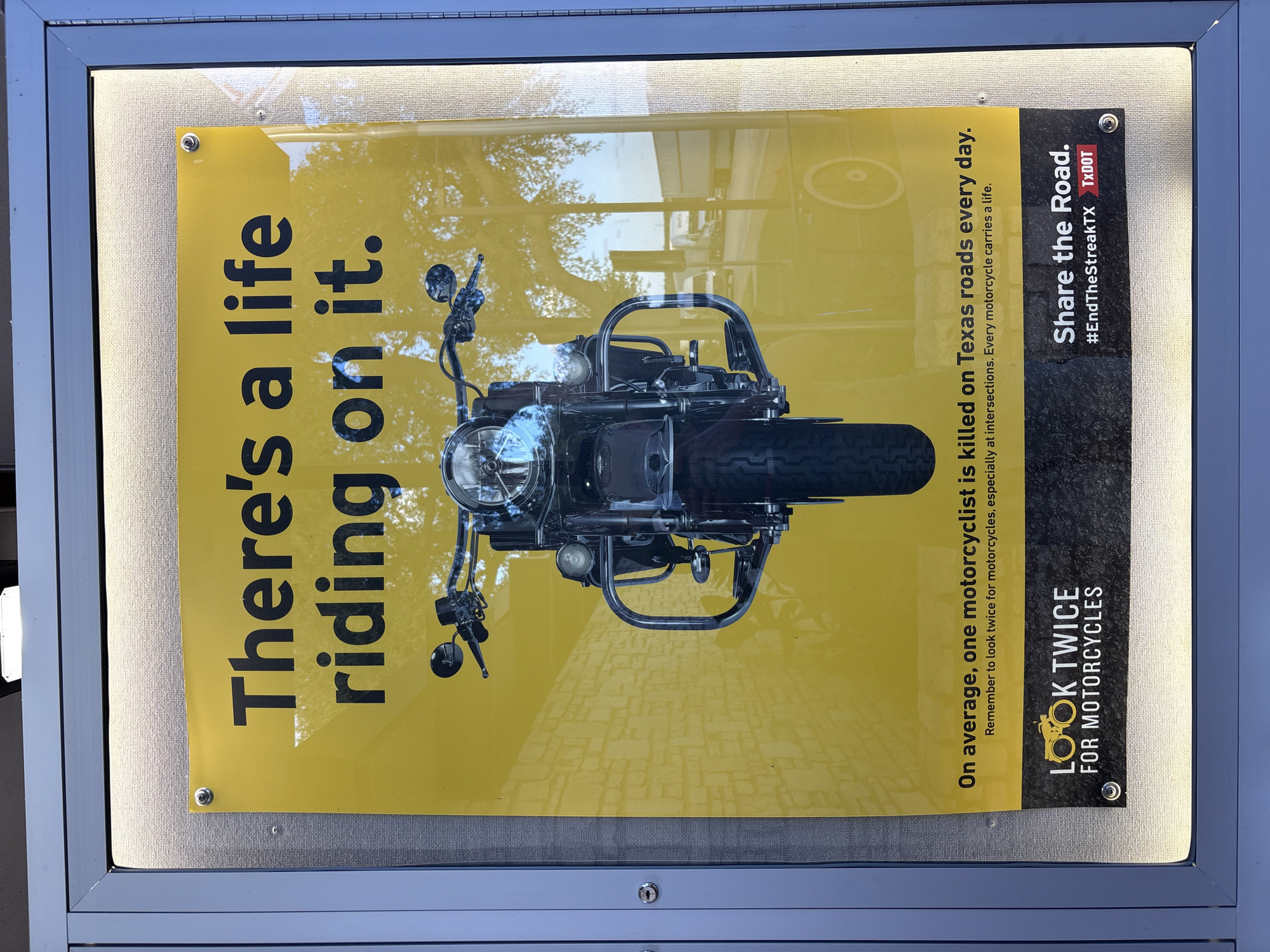 The image shows a close-up of a poster in a public display case in Sonora, United States. The poster has a yellow background and features a large image of a motorcycle in the center. The text on the poster reads: "There's a life riding on it. On average, one motorcyclist is killed on Texas roads every day. Remember to look twice for motorcycles, especially at intersections. Every motorcycle carries a life." Below this text, there is a section with two logos: "LOOK TWICE FOR MOTORCYCLES" and "Share the Road. #EndTheStreakTX TxDOT". The display case has a metal frame and is illuminated from behind. The environment appears to be outdoors, with reflections of trees and vehicles visible on the poster.