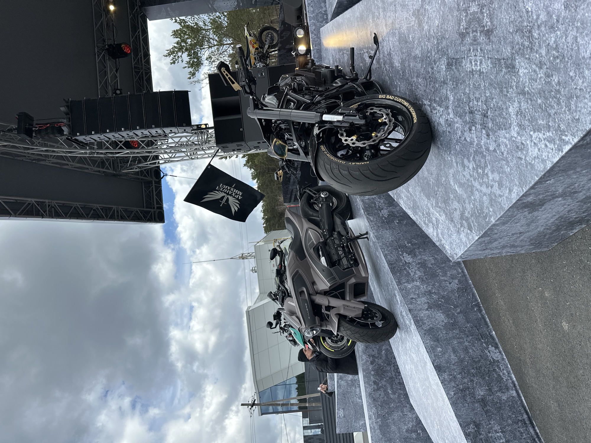 The image captures a scene in Yekaterinburg, Russia, depicting several motorcycles displayed on elevated concrete platforms. A black flag with a white design featuring an archangel figure and Cyrillic text stands prominently. The backdrop includes a stage structure with speakers and rigging. Several individuals are visible in the scene, some appearing to observe or interact with the motorcycles. A building with large glass windows is also in the background. The sky is overcast with clouds.