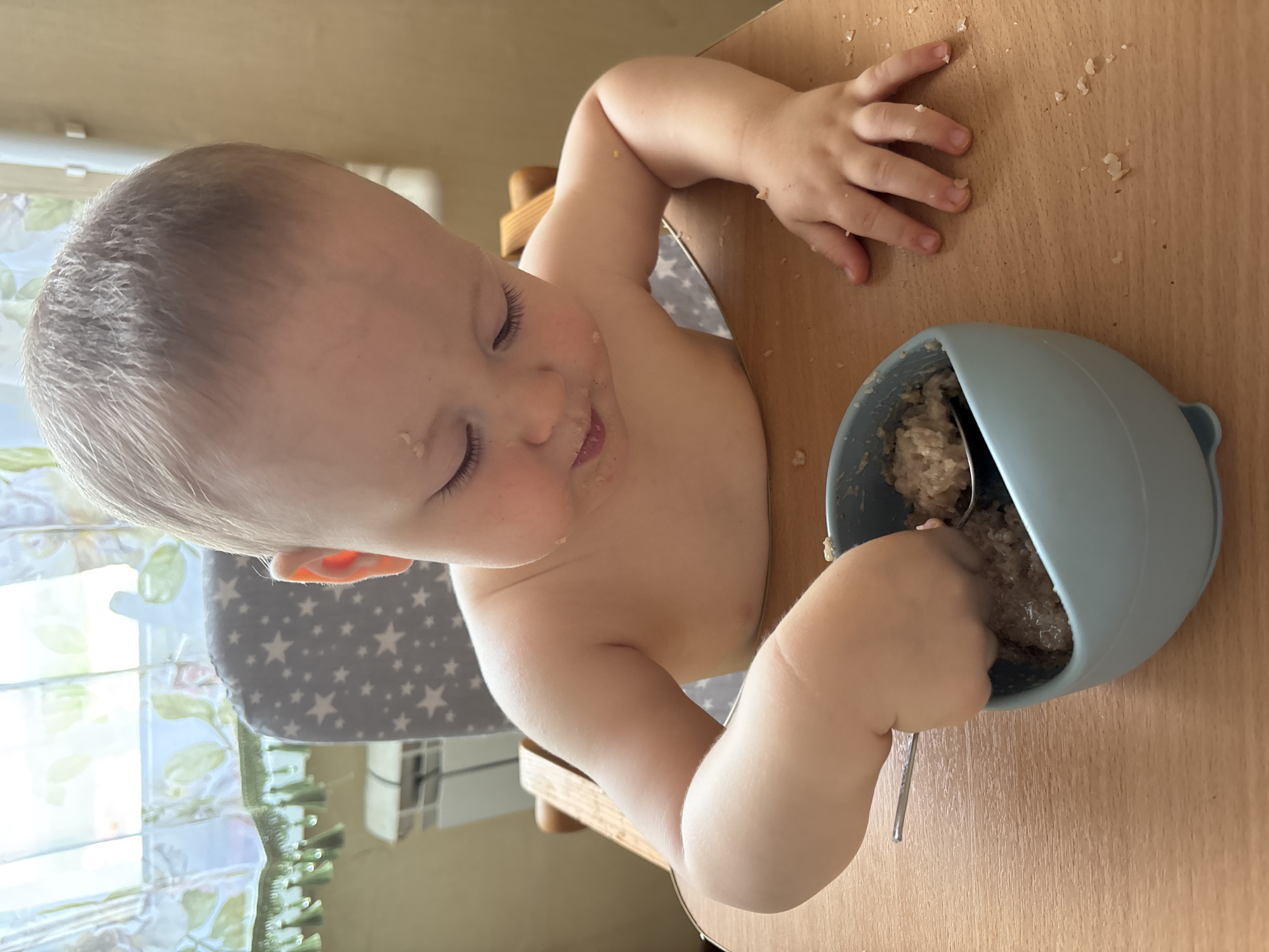 A young child is sitting at a wooden table in a high chair, eating from a blue bowl with a spoon. The child is looking down at the bowl, with food smeared on their face and chest. The high chair is gray with white stars. Behind the child, a window with sheer curtains is visible, as well as a heating radiator. The scene appears to be indoors, likely a home environment. The exact location within Naberezhnye Chelny, Russia cannot be determined from the image alone.