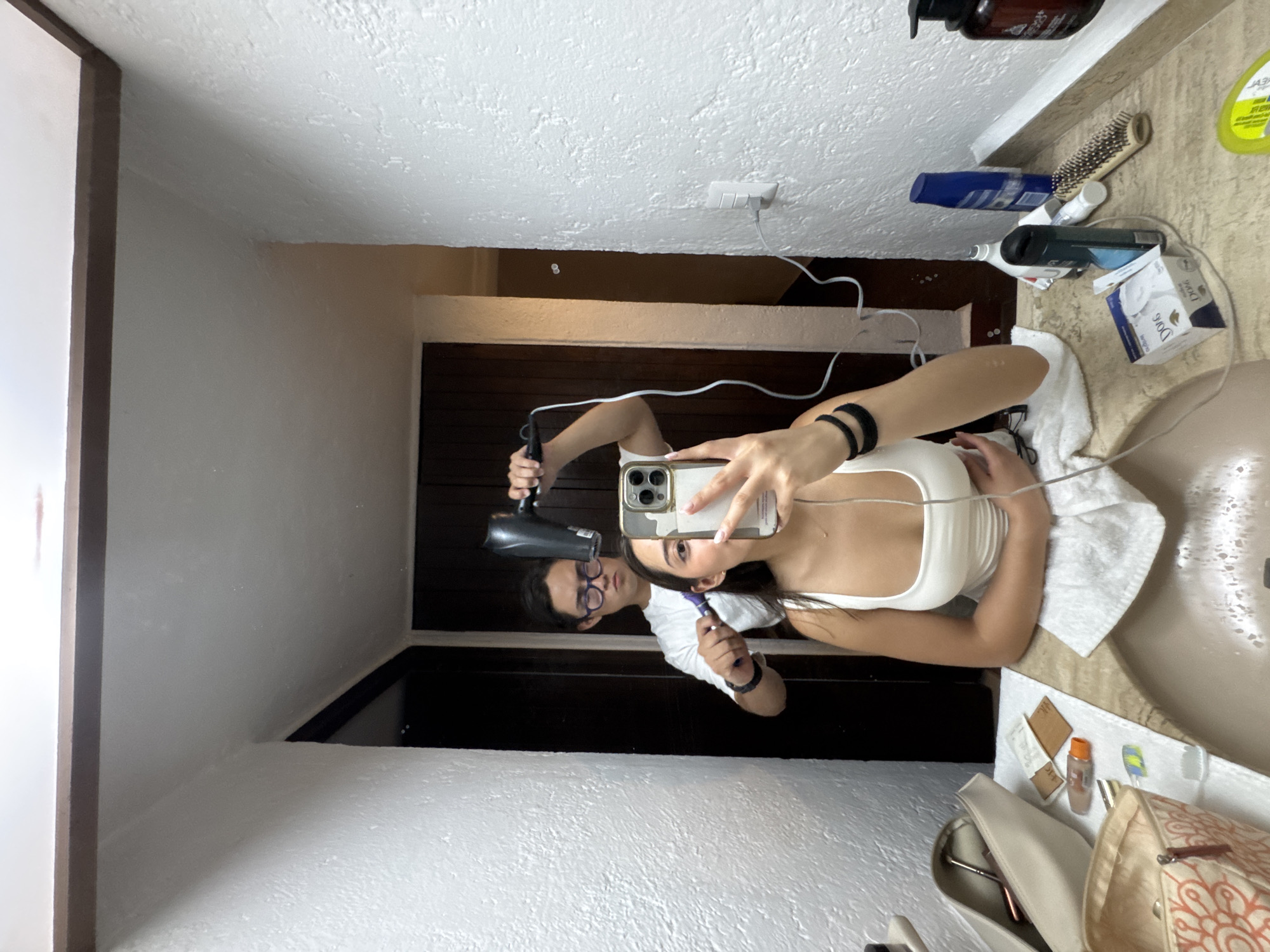 A mirror selfie shows a woman in a white tank top in a bathroom in Cocoyoc, Mexico. She holds a smartphone with two black hair ties on her left wrist, taking the picture. Behind her, a man in glasses and a white t-shirt holds a black hairdryer and comb. The bathroom has a sink with toiletries, a towel, and a bag on the countertop. The wall has a power outlet. The door frame behind the couple is dark brown wood. The room is lit with white light.