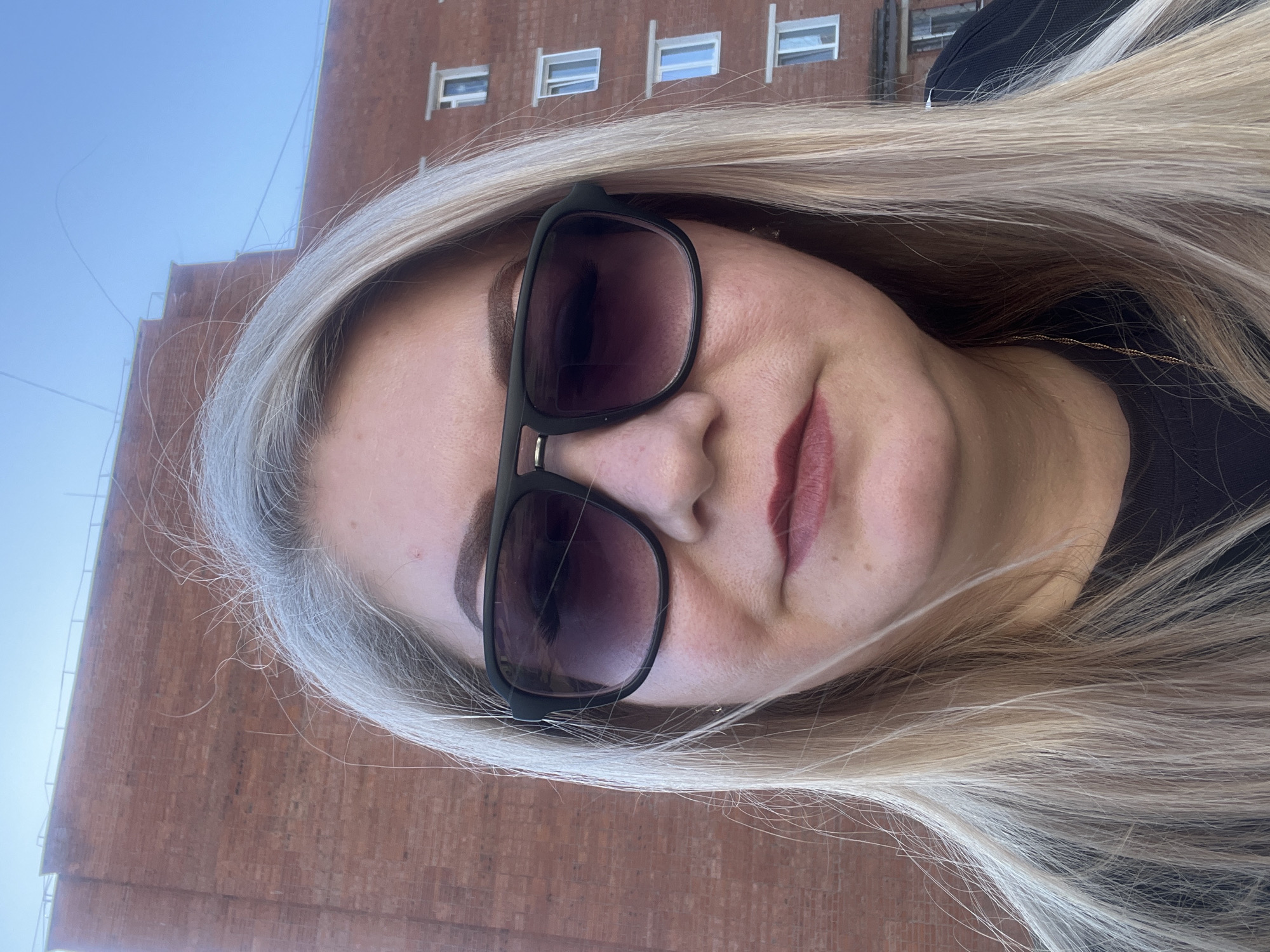 A woman with long blonde hair and sunglasses stands in front of a brick building in Glazov, Russia. The building has rectangular windows, visible in the background behind the woman. The woman's face is angled towards the camera, and she is wearing dark lipstick. A gold chain necklace is partially visible under her hair. The sky above the building is pale blue.