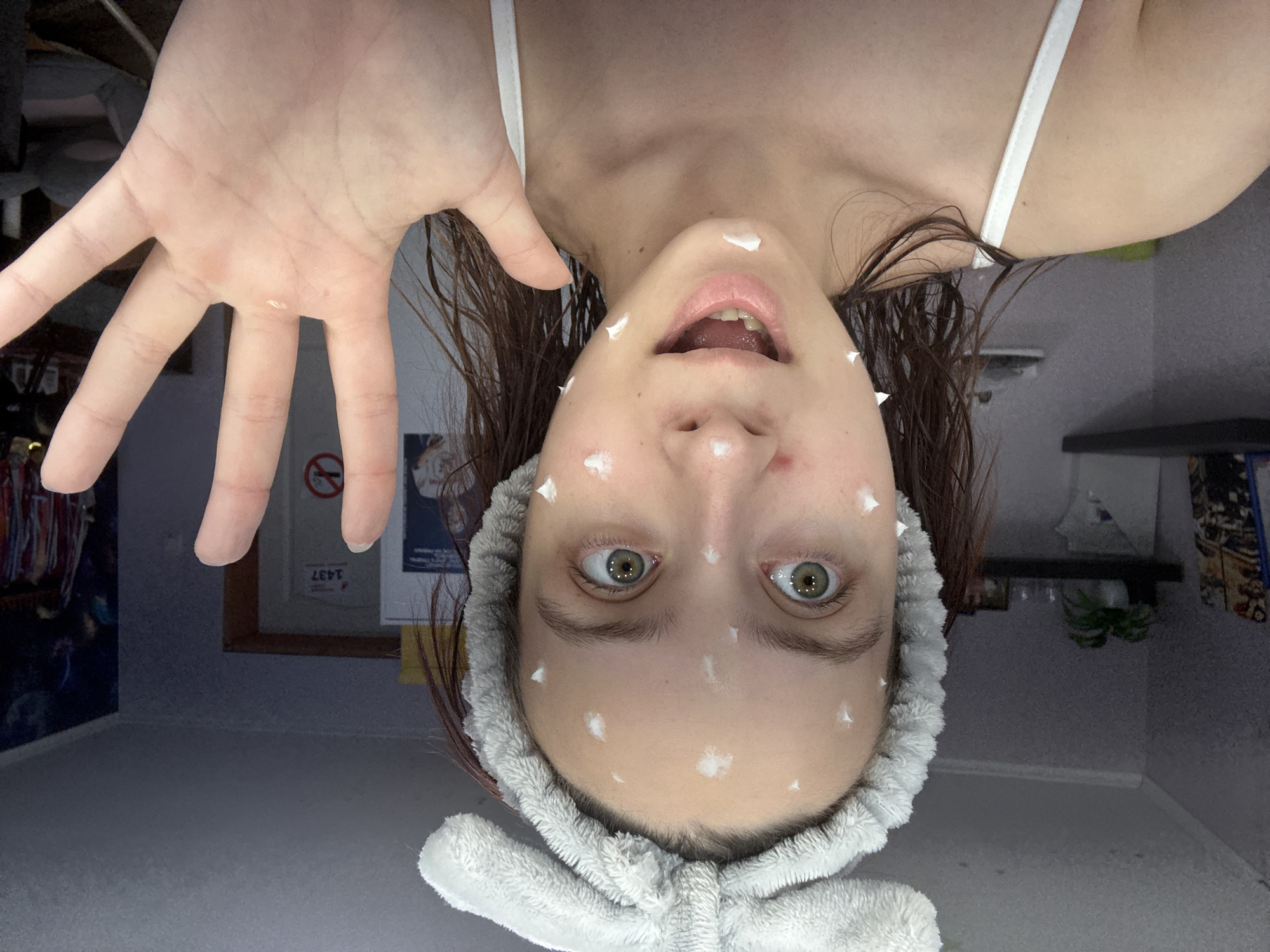 A woman with wet, brown hair and green eyes, is oriented upside down. She has white cream dots applied to her face and is wearing a light grey spa headband. Her mouth is open. She is also wearing a white tank top. Her left hand is extended towards the camera, also upside down, with a dot of cream on it.
In the background, a shelf with a plant, framed art, and a blue wall with an octopus are visible. The image appears to be taken inside a room. The location is Прикамский, Russia.