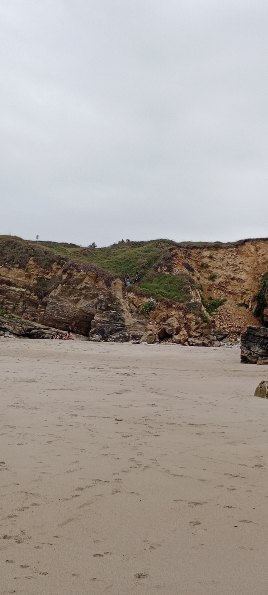 An overcast day at a beach in an unknown location in Spain shows a wide expanse of light brown sand in the foreground. In the middle ground, a rocky cliff face with patches of green grass and vegetation rises up. A group of people are visible on the beach, some sitting together in the shade of the cliff, while others are standing on top of the cliff, looking out. The sky is a uniform pale gray, with no visible sun. The scene suggests a quiet, somewhat overcast day at the beach.