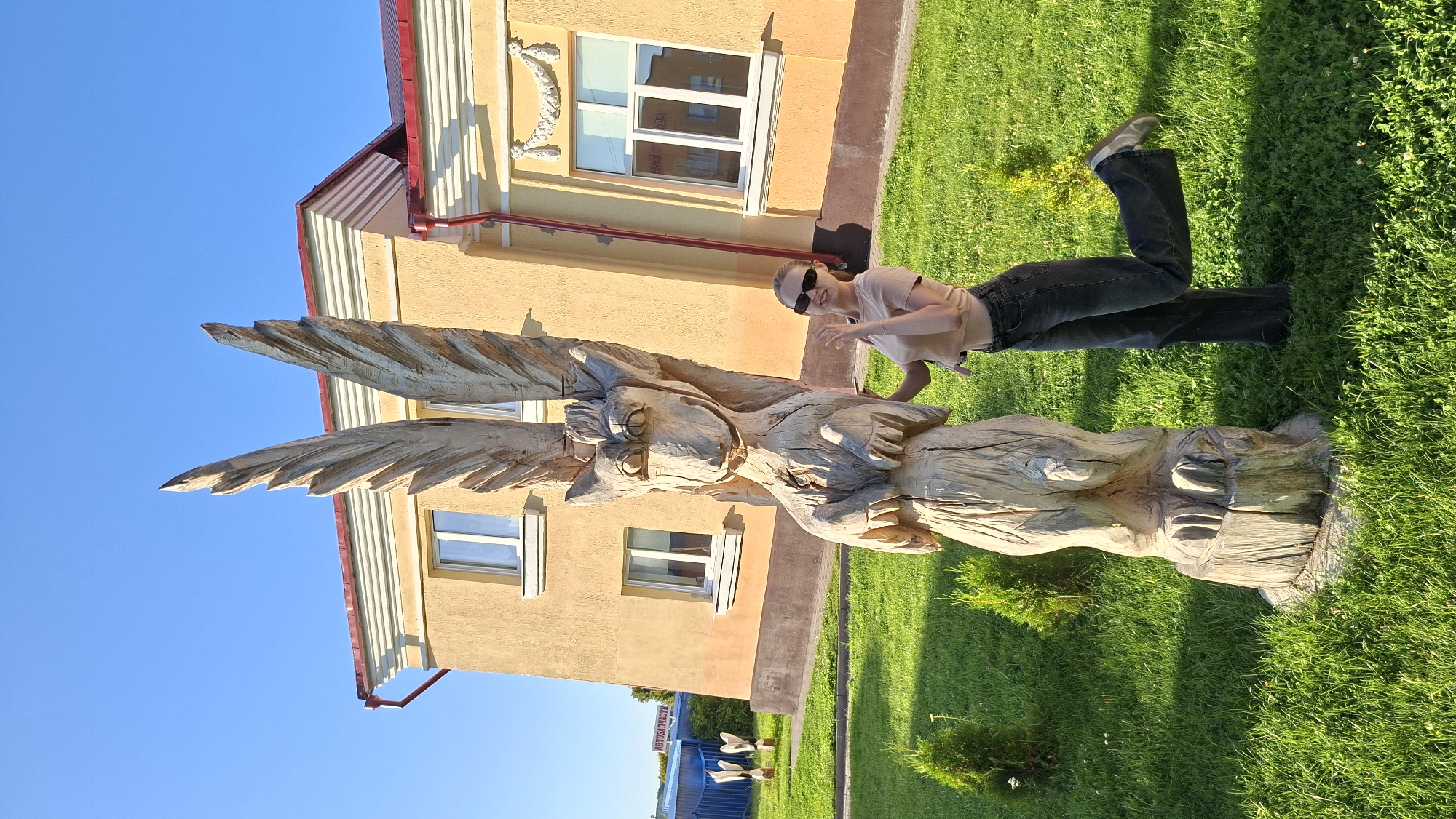 A young woman with blonde hair and sunglasses stands on a green lawn next to a tall, wooden sculpture of a creature with wings and large eyes. The sculpture is unpainted and appears to be made from a single log. The woman is wearing a light pink t-shirt and dark wash, wide-leg jeans. She has her left leg bent and extended backward, and her right hand is raised near her face, with her fingers spread. Behind the sculpture and the woman is a light yellow building with a red roof and white-framed windows. The sky above is clear and blue. The scene is illuminated by bright sunlight, casting shadows on the grass. In the background, there are some small evergreen bushes and a blue fence with a sign that reads "АВТОЗАПЧАСТИ" (Auto Parts in Russian). The setting appears to be an outdoor area, possibly a park or the grounds of a public building, in the city of Myadzyel, Belarus.