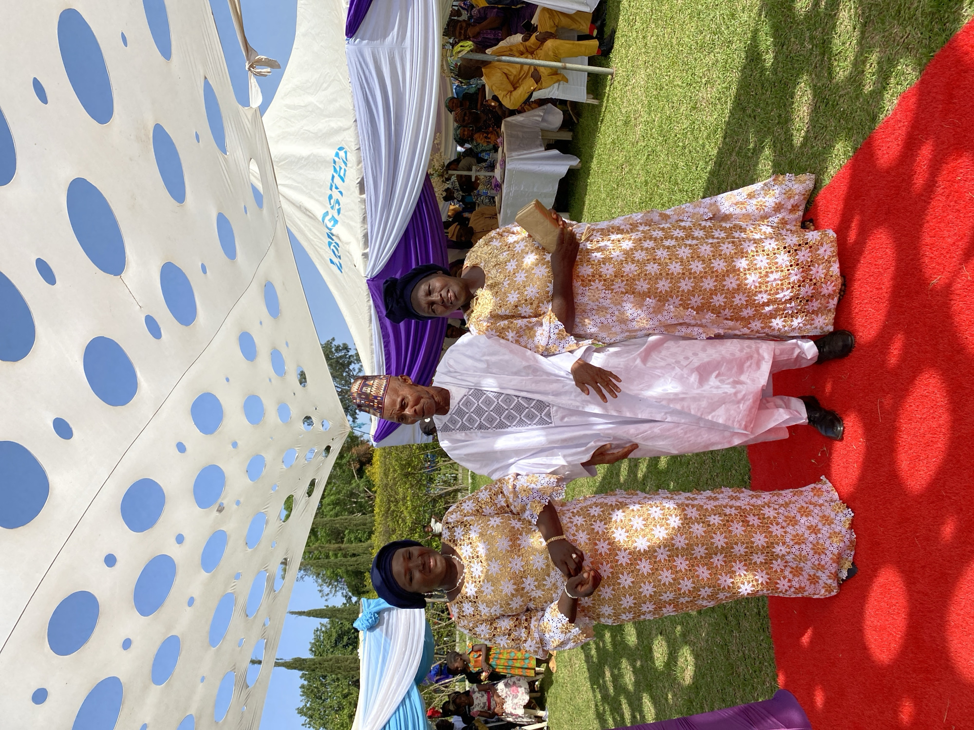 The image shows three people standing on a red carpet in an outdoor setting in Bukuru, Nigeria. Two women, dressed in matching gold and white floral lace dresses and blue headwraps, flank a man in the middle. The man is wearing a white traditional Nigerian outfit with a patterned cap. Behind them are white and purple draped tents, and some additional people seated under the tents. The ground is covered with grass, except for the red carpet. The sky is visible through holes in the tent.