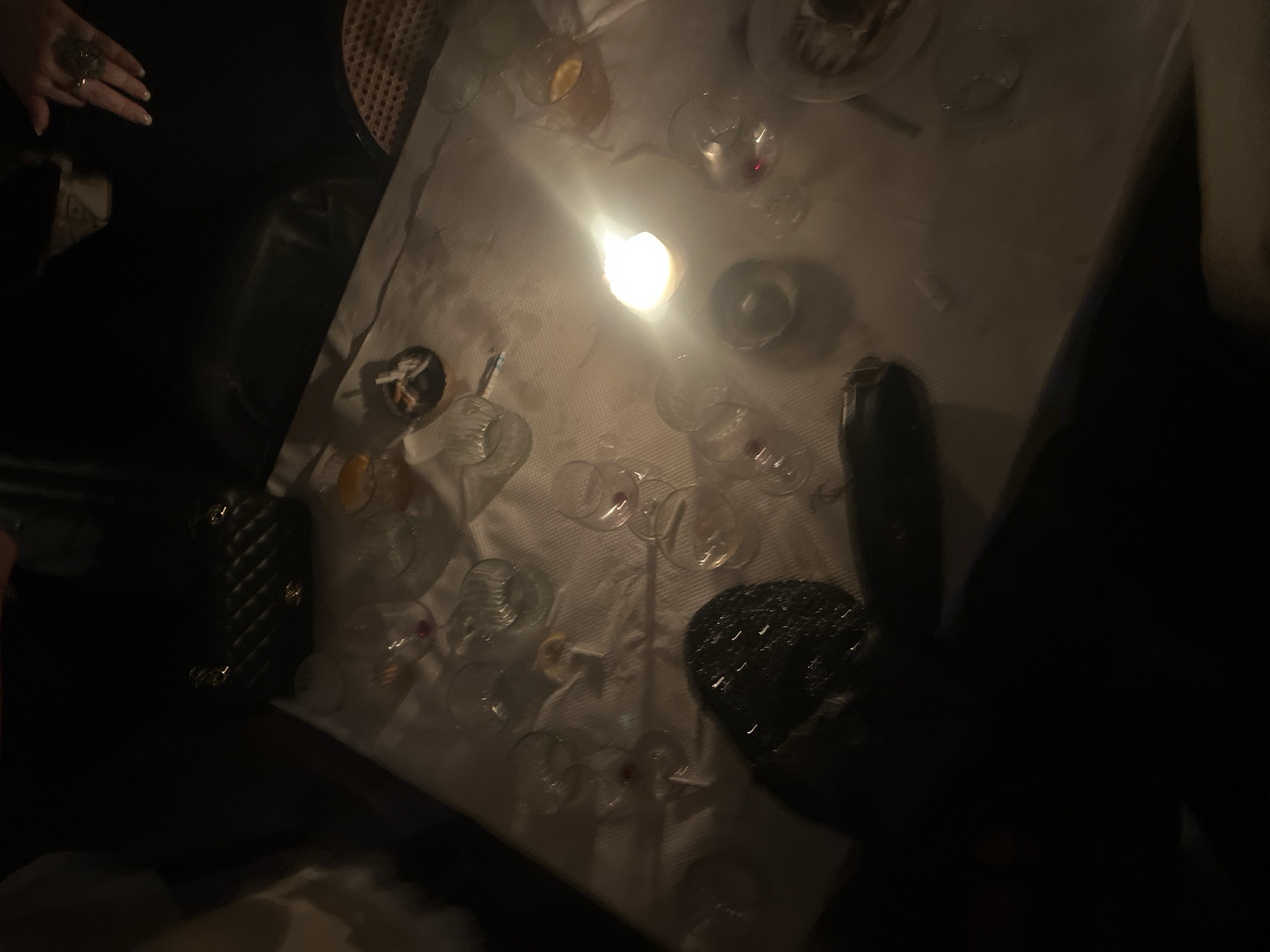 The image shows a dimly lit dining table in Paris, France, covered with a white tablecloth. Several wine glasses and some filled with liquid are scattered across the table. A lit candle provides the main source of illumination. There are also plates and cutlery on the table. A purse or bag is placed on a chair near the table. A woman's hand with a ring is visible at the top left. There are people seated around the table, partially visible on the right and bottom of the image. The setting suggests a restaurant or private dining space.