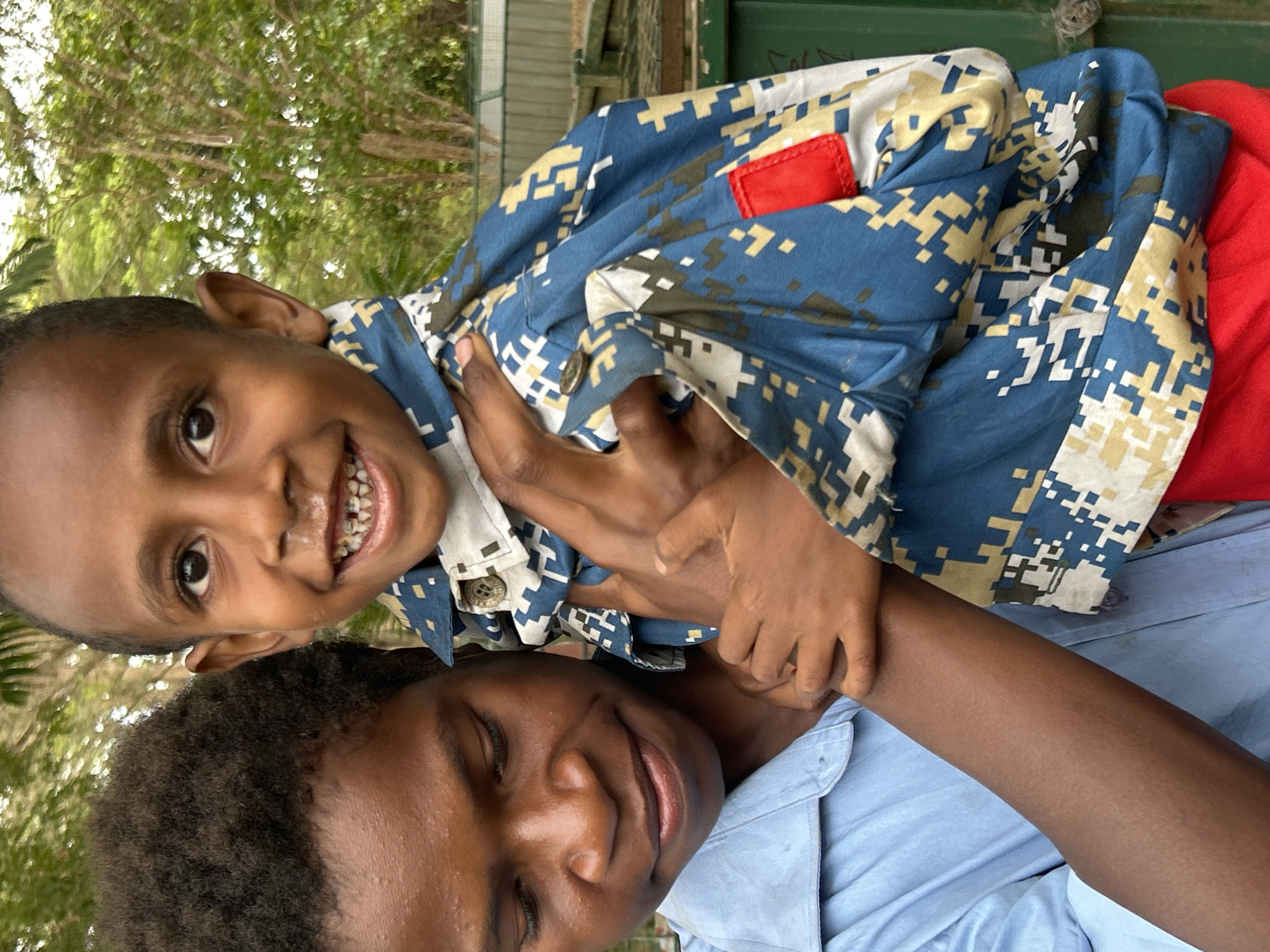 Two individuals are depicted in a close-up, outdoor shot. The foreground is dominated by the head and upper torso of a younger child, held by an older individual. The younger child, facing the camera, has dark skin, black eyes, and a wide smile revealing teeth with visible decay. They are wearing a blue shirt with a digital camouflage pattern in blue, white, beige, and green. A small red tag is visible on the shirt's shoulder.
Behind and to the left of the younger child is the face and shoulder of the older individual, who is looking down and to the left. This individual also has dark skin and is wearing a light blue collared shirt. Their arm is supporting the younger child.
The background suggests an outdoor environment with lush green foliage, likely trees, creating a soft, blurred bokeh effect. A green structure, possibly a dumpster or bin, is visible on the right side of the frame. A fence, likely chain-link, is also present in the mid-ground. The lighting indicates a bright, possibly sunny day. The scene appears to be set in a residential or community area within Tanubada, Papua New Guinea, given the tropical vegetation and the individuals' likely ethnic background. The interaction between the two suggests a caring relationship, possibly sibling or parent-child.