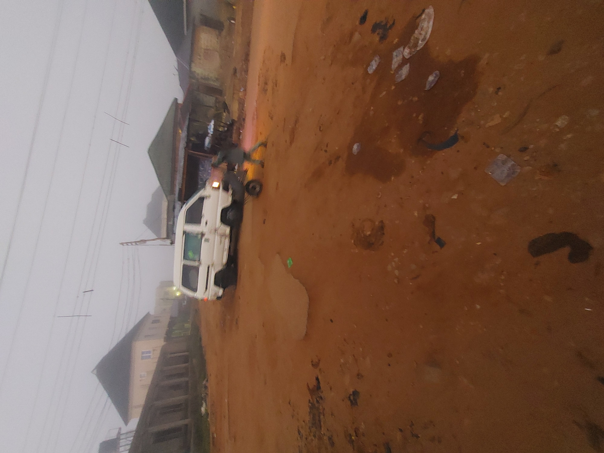 In Bukuru, Nigeria, a white van is parked on a dirt road. A person is pushing a wheelbarrow alongside the van. The environment appears to be a rural or semi-urban area with some buildings in the background. The sky is overcast, suggesting a cloudy or foggy day. The ground is composed of reddish-brown dirt, with some scattered debris and puddles visible. The overall scene depicts everyday life in this Nigerian location.