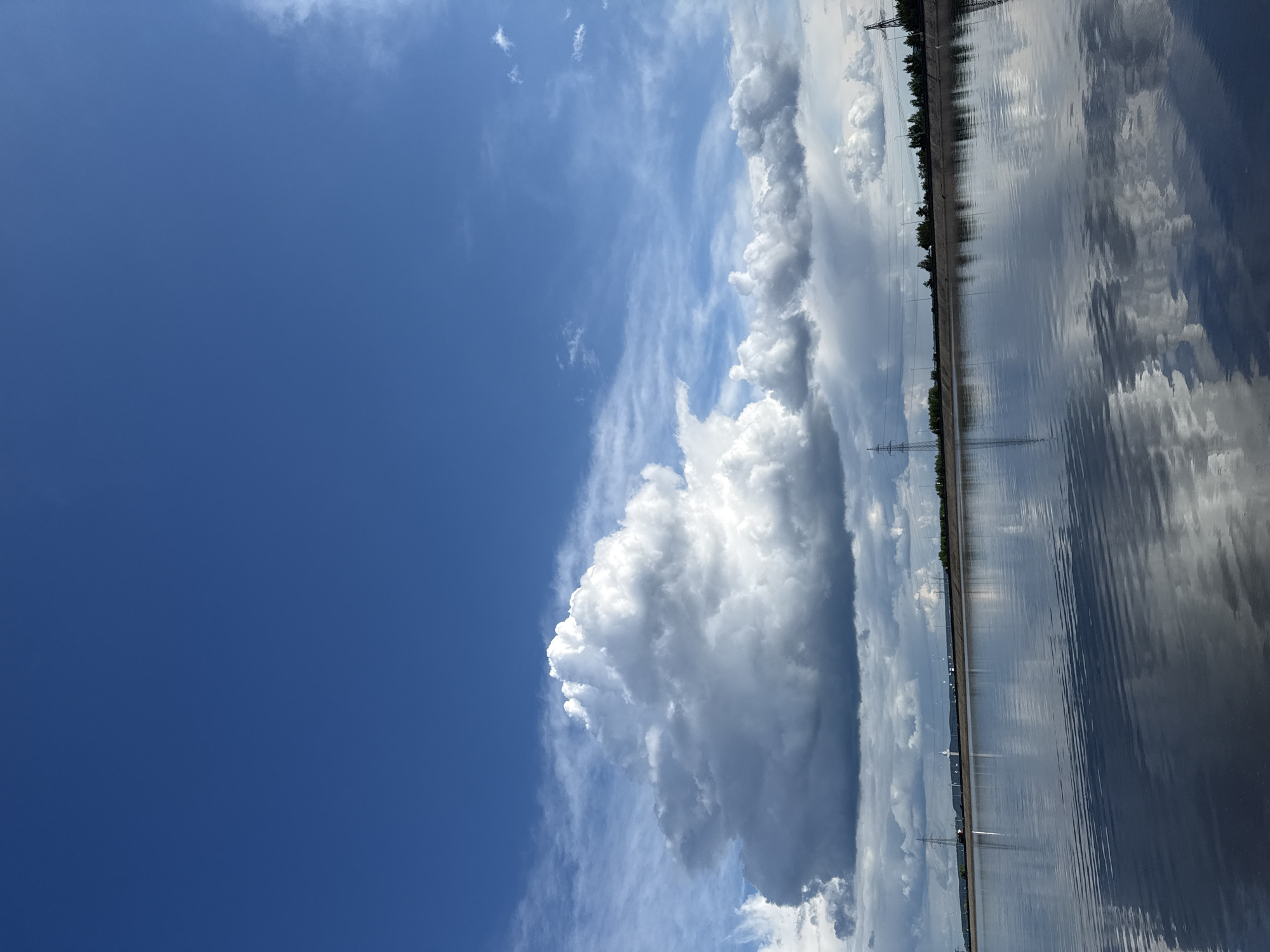 The image shows a landscape in Noviy, Russia. The sky dominates the left side of the frame, transitioning from a clear, deep blue to wispy clouds and a large, towering cumulonimbus cloud formation. On the right side of the image, a long body of water, likely a river or reservoir, reflects the clouds and the sky above. Along the far edge of the water, there is a line of trees and vegetation, interrupted by utility poles and structures. The reflective surface of the water and the placement of these features suggest a flat, expansive landscape.