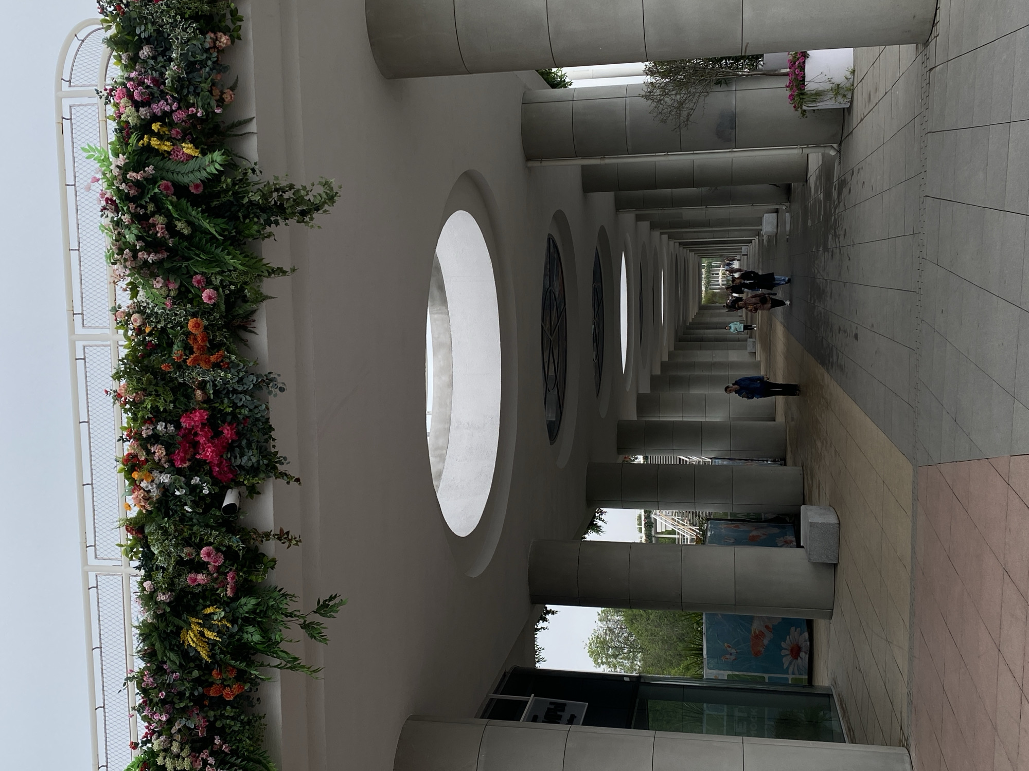 The image captures a perspective view of a colonnade located in an urban environment. The structure consists of a row of evenly spaced, cylindrical columns that support a vaulted ceiling with circular windows. Some of the windows feature decorative stained glass. The walkway is paved with varying shades of grey, beige, and red tiles.
Along one side of the structure, a dense vertical garden of mixed flowers and foliage adds a burst of color to the otherwise neutral palette of the architecture. The garden appears to be artificial.
Several individuals are walking through the colonnade.
The scene suggests a public space, possibly a pedestrian thoroughfare.