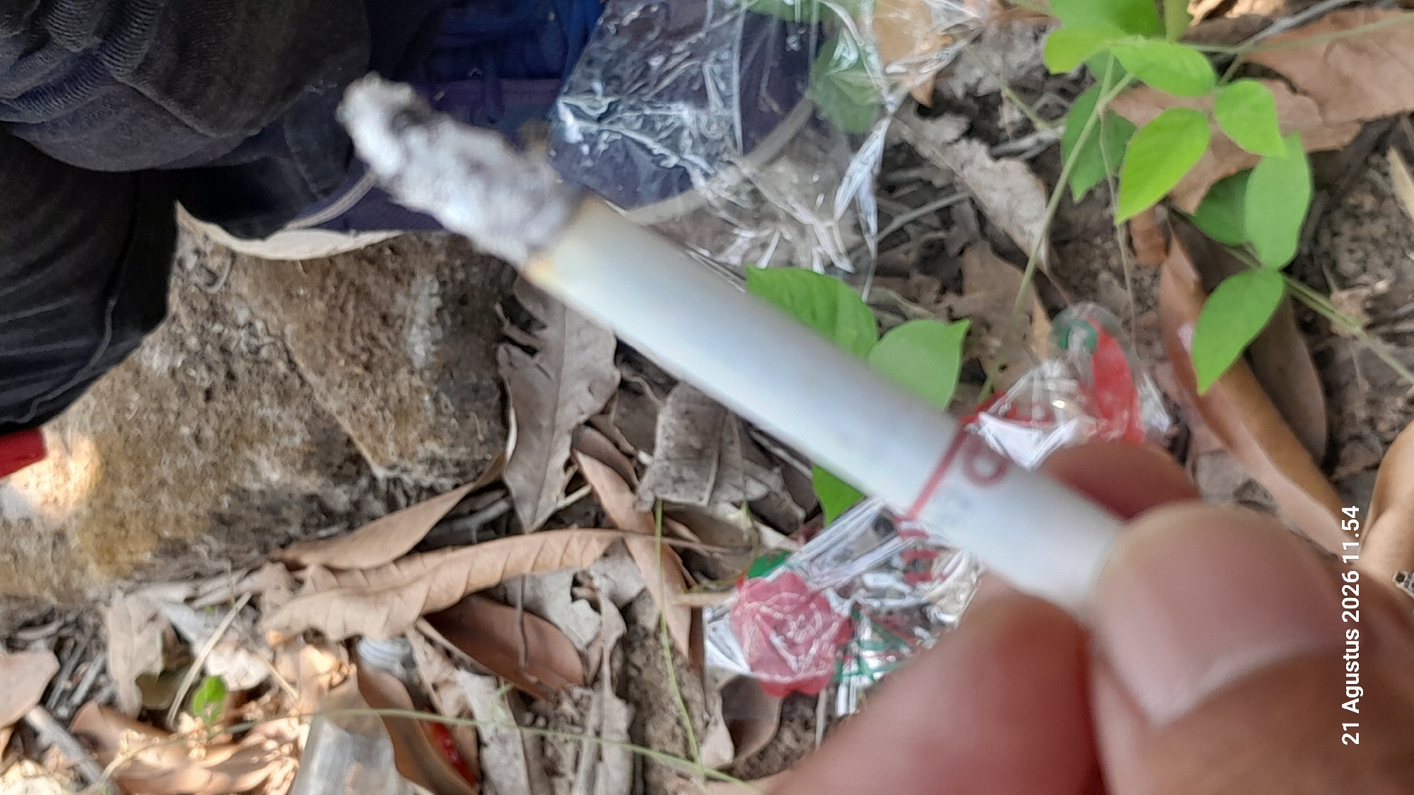 A close-up, over-the-shoulder shot shows a person holding a lit cigarette between their fingers. The cigarette is white with a red band near the filter. The background consists of dry leaves, dirt, and some green foliage, suggesting an outdoor environment, possibly in Takeran, Indonesia. Several discarded plastic wrappers, one with red and silver accents, are visible on the ground. The person's hand is in the foreground, obscuring part of the scene. The timestamp "21 Agustus 2026 11.54" is overlaid on the bottom left of the image.