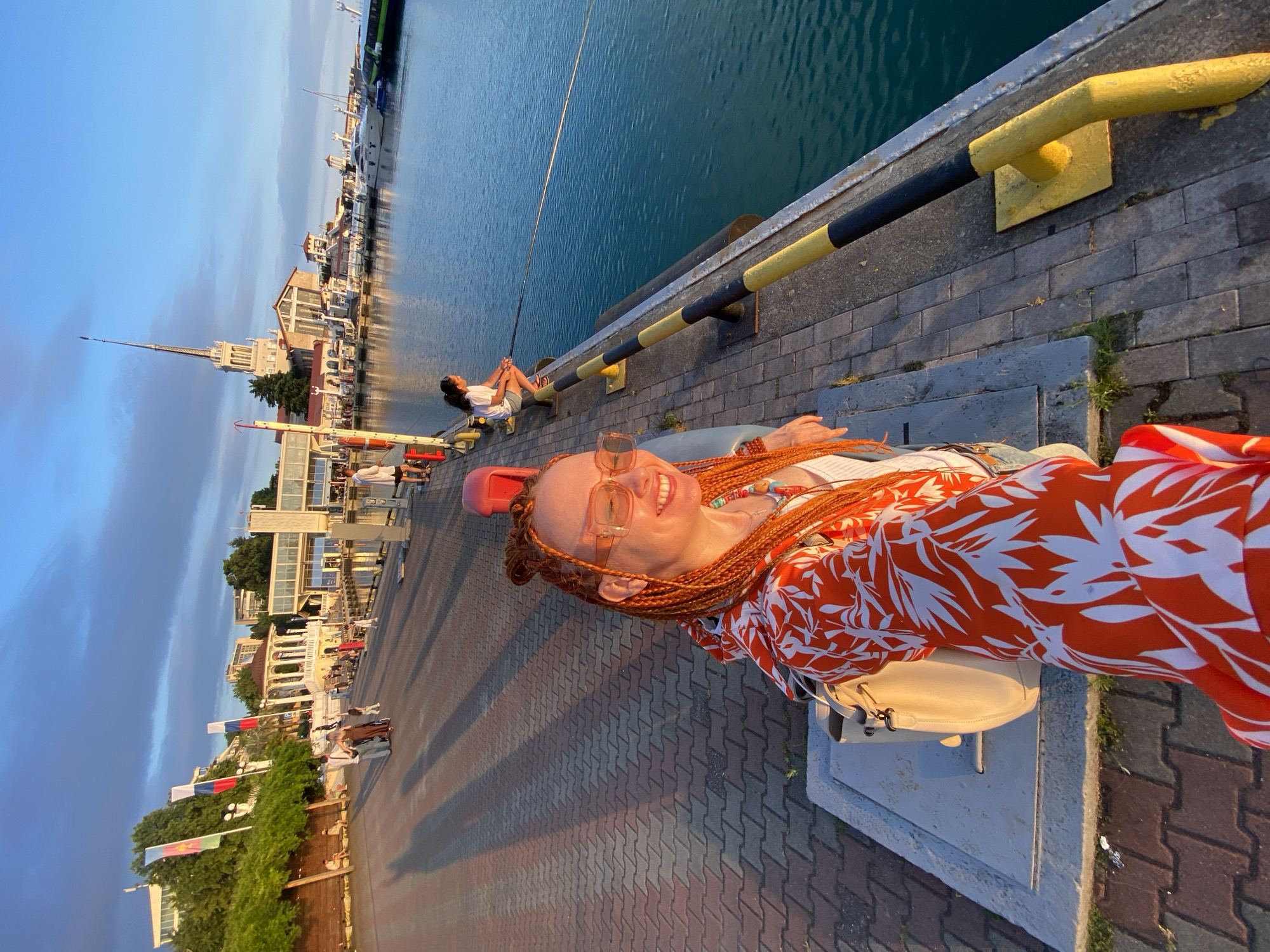 The image shows a woman with long, braided auburn hair and glasses, wearing an orange and white floral print dress. She is smiling and lying down on a concrete structure along a waterfront promenade. Behind her, a person with dark hair is sitting with their legs crossed, looking out at the water. In the background, there are buildings with white facades and arches, flags flying, and boats docked in the harbor. The sun appears to be setting, casting long shadows across the brick-patterned pavement and illuminating the scene with warm light. The sky is a deep blue with streaks of white clouds. The overall atmosphere is relaxed and summery. The location is identified as Sochi, Russia.