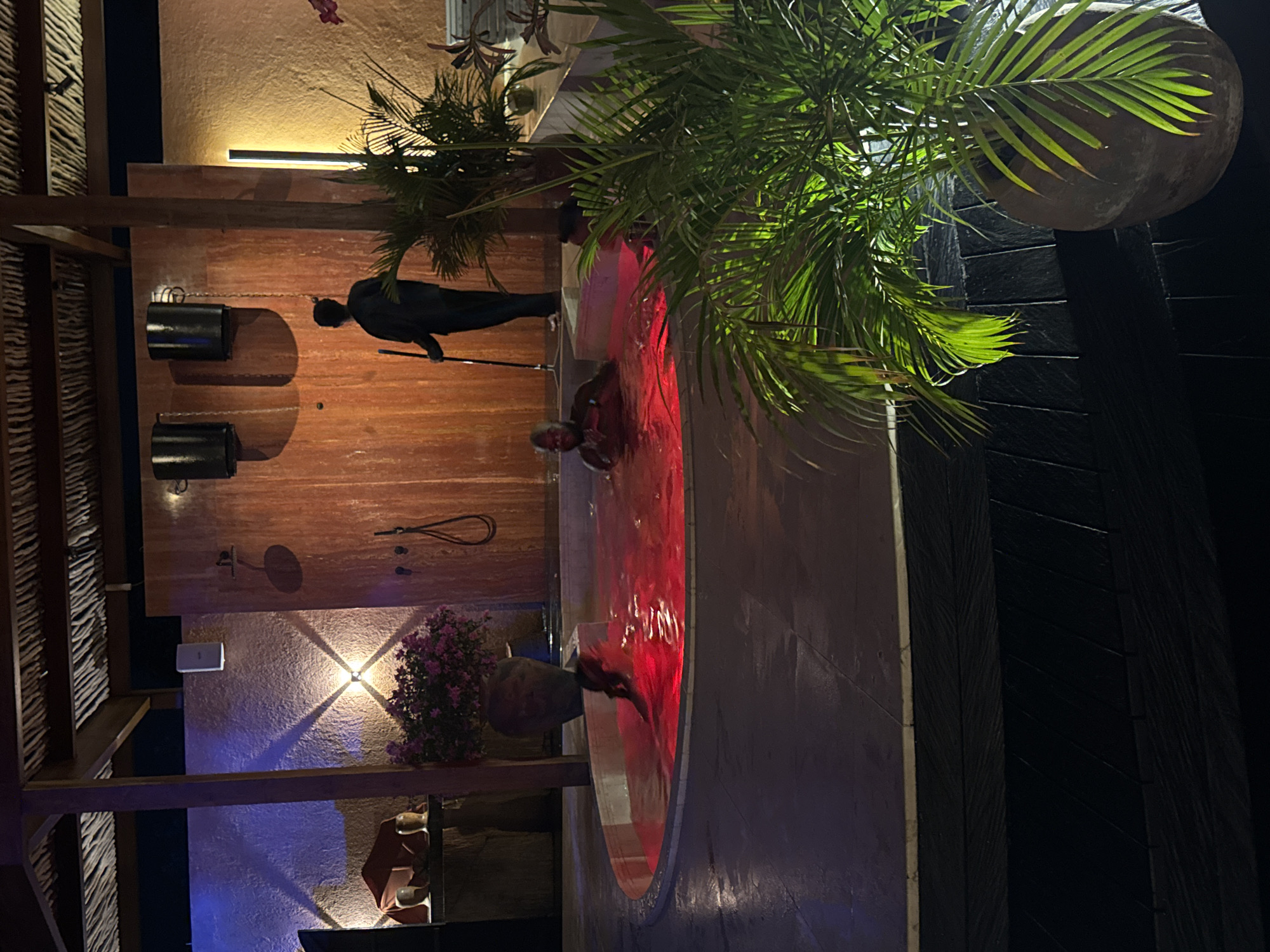 The scene depicts a semi-enclosed, dimly lit leisure area at night, located in Kuta Selatan, Indonesia. A large, circular hot tub with internally lit red water occupies the lower-middle ground. Two individuals are partially submerged in the hot tub; one adult male with dark skin and a bald head is visible from the chest up on the right side, facing left, while another individual is partially visible on the left side, mostly obscured by the water.
Behind the hot tub, an adult figure, silhouetted against a reddish-brown wooden wall, stands upright, holding a long dark pole. This figure is facing away from the viewer. Above this figure, two dark cylindrical objects resembling buckets or large sconces are mounted high on the wooden wall, casting prominent shadows below them. To the right of the standing figure, a vertical strip light emits a warm glow. Further right, a large potted plant with green palm-like leaves partially obscures the bottom right foreground.
To the left of the hot tub, a textured, light-colored wall features a white rectangular object mounted high, possibly a speaker. Below it, a warm-toned wall sconce casts distinctive X-shaped light patterns and shadows. Further left, a subtle blue uplight illuminates a section of the wall. A large dark pot containing pink flowering plants is situated on a raised platform near the hot tub's edge. To the left of these flowers, two golden, ornate vessels are placed on a dark surface. The ceiling above is constructed from natural, woven material supported by dark wooden beams. The flooring around the hot tub is a dark, tiled surface.