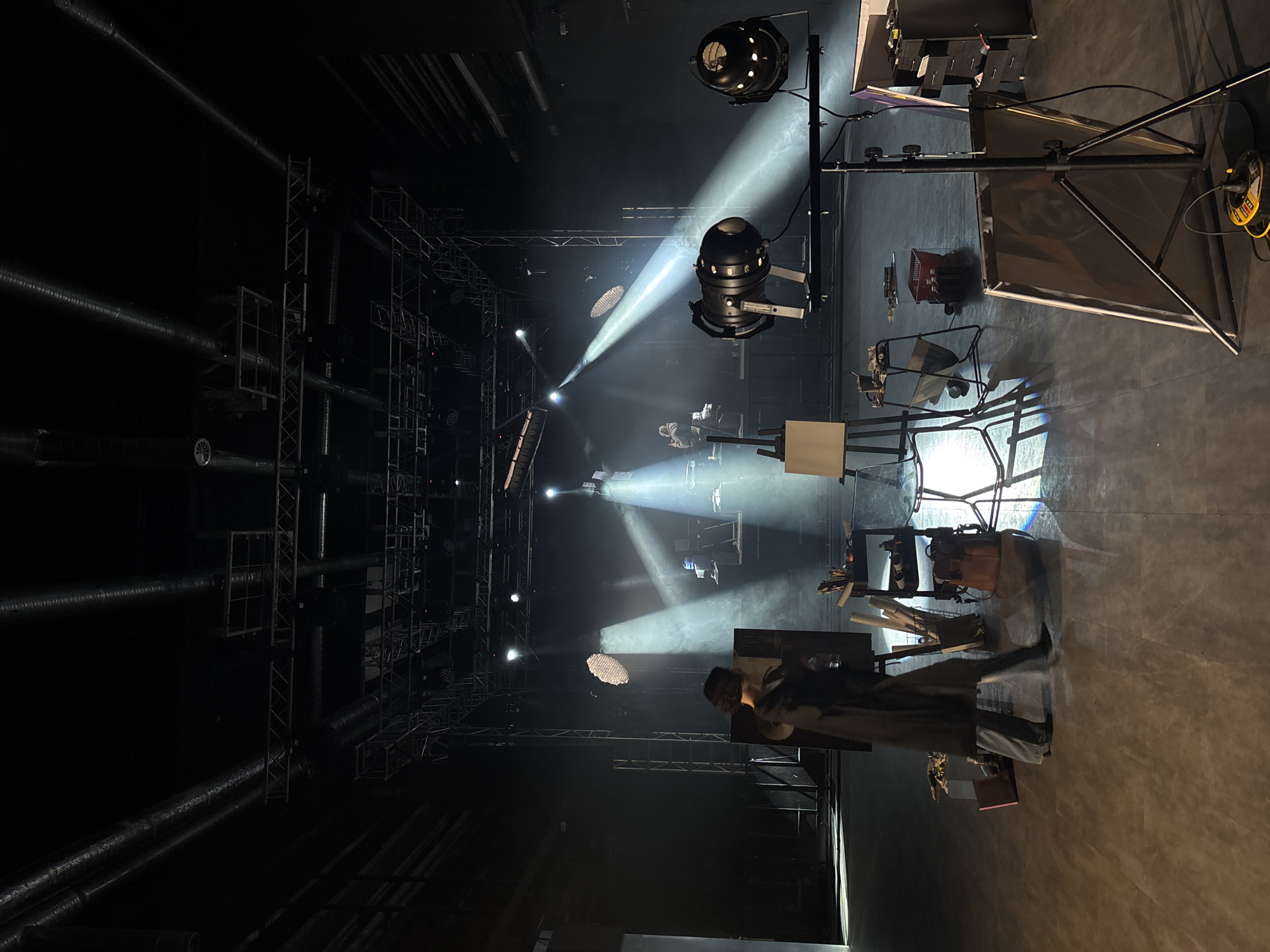 The image depicts a dark, expansive indoor space, strongly resembling a theatre stage, concert hall, or large production studio. The environment is illuminated by dramatic, focused beams of artificial light cutting through what appears to be a hazy atmosphere.
In the immediate foreground, to the right, a person is partially visible, standing near several pieces of equipment. This includes a tall black stand supporting a large, rectangular diffuser or reflector panel. Nearby, a wooden easel stands upright, and a small cart or table is laden with various items like bags, papers, bottles, and brushes. The floor is a dark, smooth surface, reflecting some of the intense light.
The midground showcases multiple stage lighting fixtures, including spotlights and other luminaires, mounted on stands and extensive overhead metal truss structures. Strong white light beams originate from these fixtures, creating distinct patterns on the stage and illuminating specific areas. Another person is visible further back in one of these lit zones.
The background and overhead areas reveal a complex network of metal trussing, support beams, and large ventilation pipes, common in professional performance venues. The overall scene suggests a technical rehearsal, setup for an event, or a photoshoot/videography session, rather than a live performance, given the visible equipment and sparse presence of people.
The time of day is indiscernible as the scene relies entirely on artificial lighting. No specific emotions are conveyed, as the focus is on technical setup. While some labels might be present on the equipment, no clear, legible text is visible. The location is Moscow, Russia.