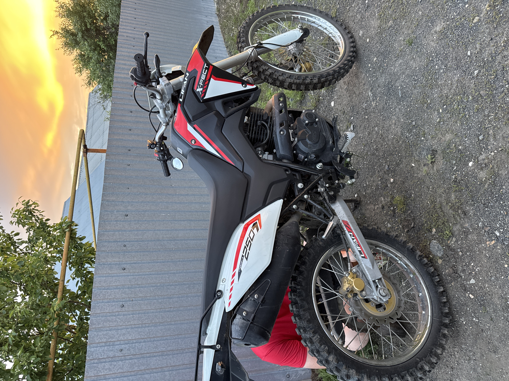 A dirt bike, a Lifan X-Pect 250, is parked outdoors on a gravel surface in front of a corrugated metal fence. The motorcycle is black and red with white accents. A person wearing a red shirt is partially visible behind the rear wheel of the motorcycle, appearing to be working on it. The background shows a tree and a glimpse of a colorful sunset sky with yellow and orange hues. The location is identified as Starokhalilovo, Russia.