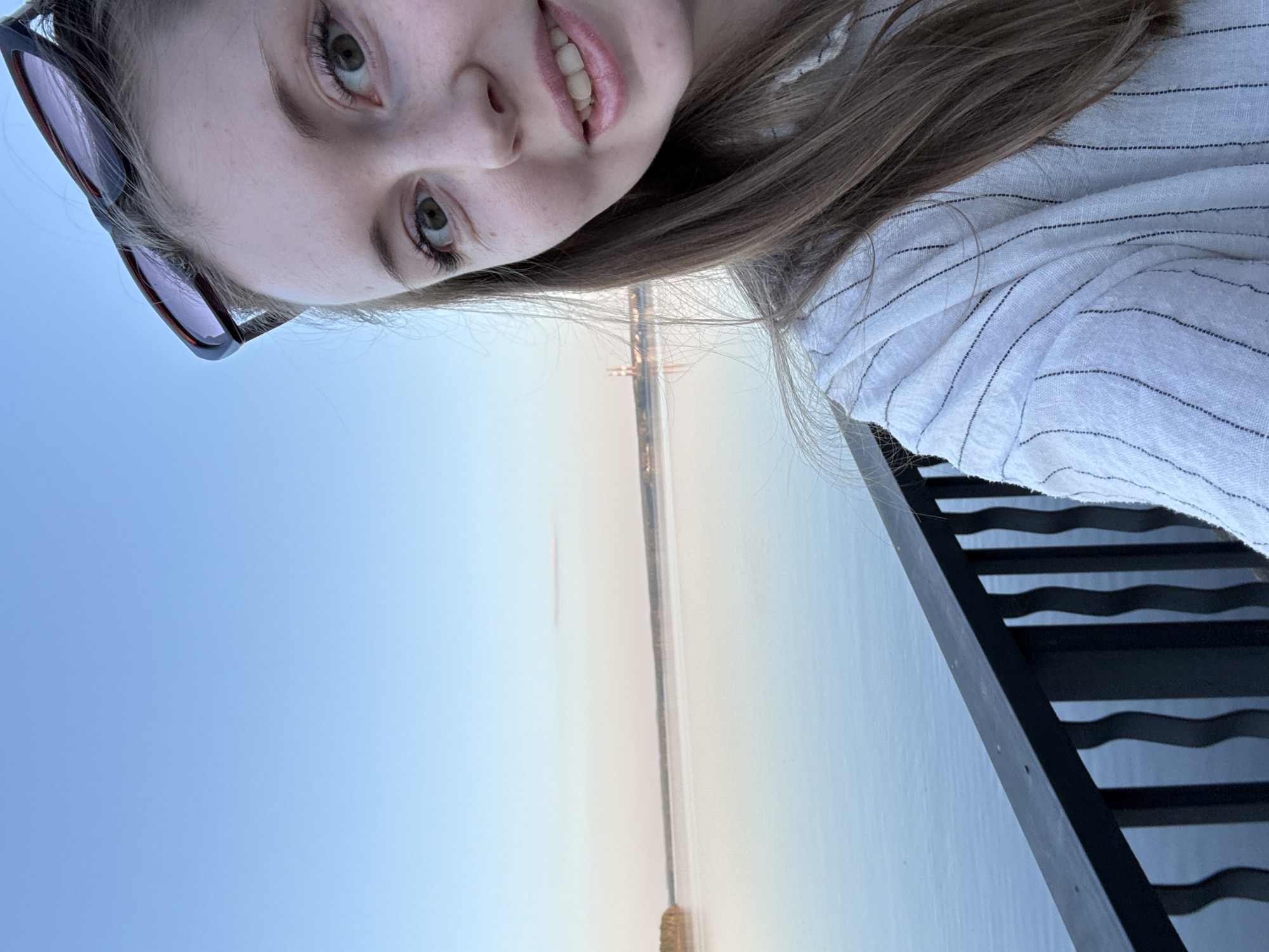 A close-up selfie shows a young woman with fair skin, light brown hair, and green eyes. She is wearing sunglasses perched on her head and a white blouse with thin, dark stripes. The background reveals a river under a clear, pale blue sky, with a bridge visible in the distance. A section of a black metal railing is visible at the edge of the frame. The location is the city of Unknown, Unknown.