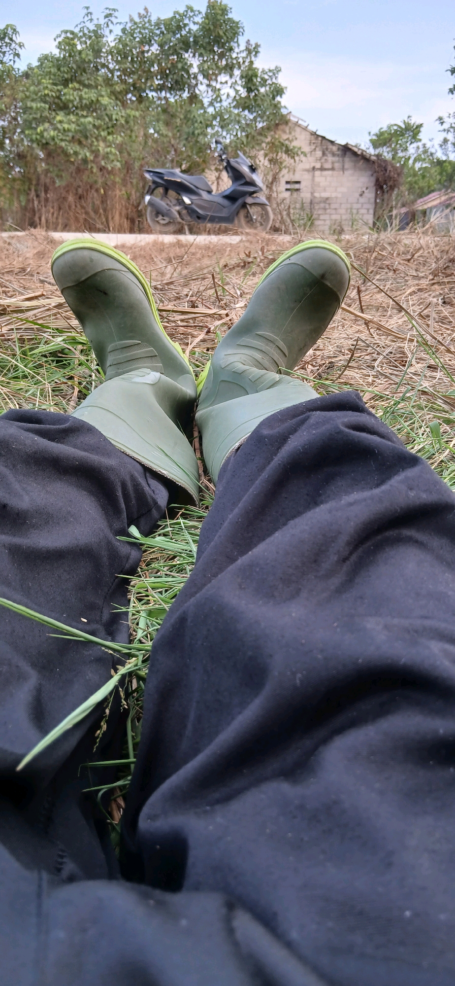 The image captures a low-angle view of a person's legs and feet clad in dark pants and green rubber boots, resting on dry grass and some sparse green vegetation. In the background, a dark-colored motorcycle is parked near a dilapidated brick building with a partially collapsed roof. Trees with dense foliage and some dry brush are also visible. The sky appears overcast. The scene suggests an outdoor environment, possibly a rural or semi-urban area in Samarinda, Indonesia, characterized by dry conditions and basic infrastructure.