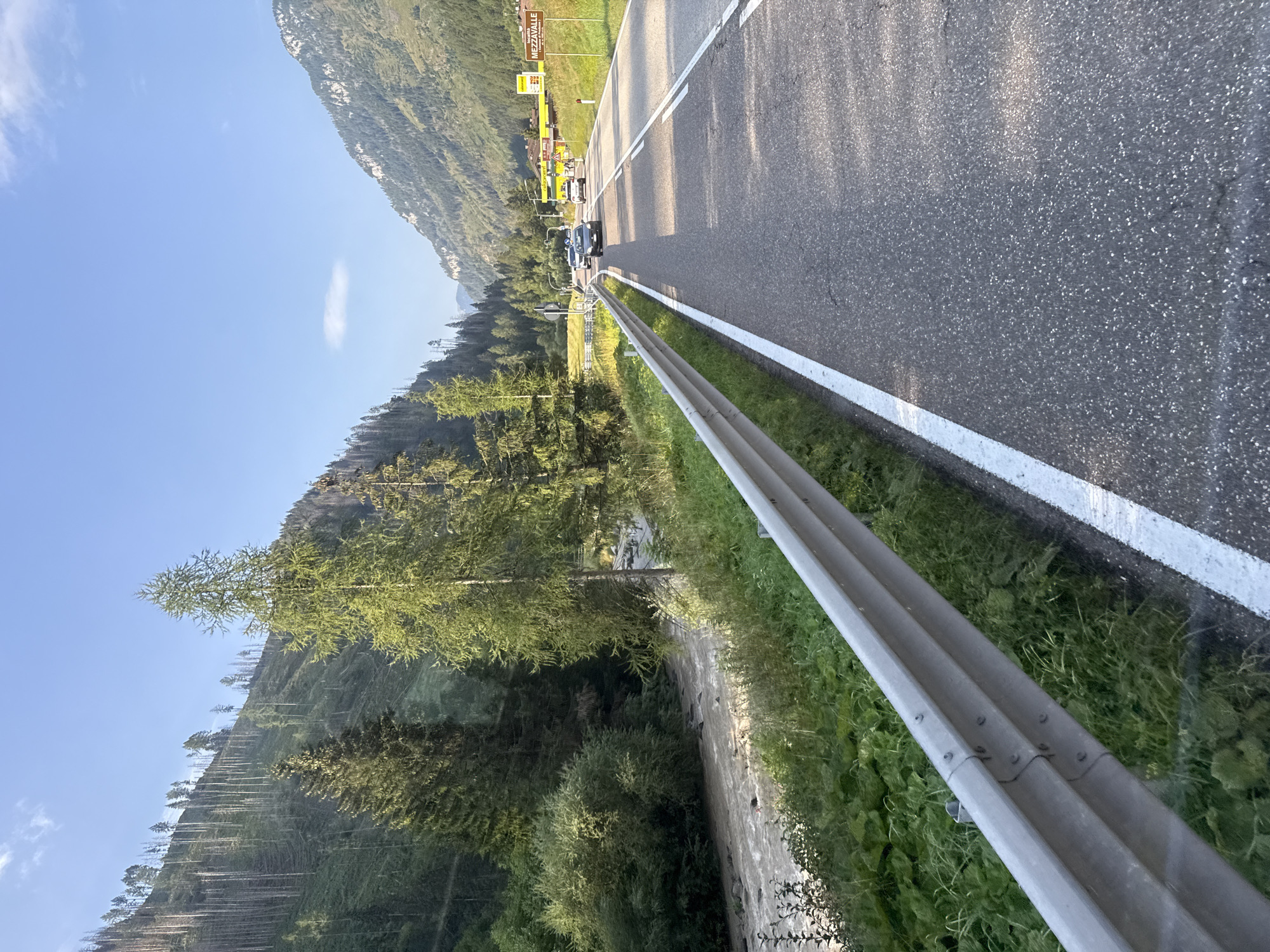 The image depicts a scenic mountain road in Italy, specifically near Mezzavalle, a location within the municipality of Predazzo. The road is asphalt, with a white line marking the edge of the lane. A metal guardrail runs along the left side of the road, separating it from a vegetated embankment and a flowing river.
The river, appearing muddy and turbulent, rushes through a rocky bed, bordered by lush green foliage and trees. On the right side of the road, a sign indicates "MEZZAVALLE". Beyond the road, a hilly landscape unfolds, covered with dense evergreen trees and patches of bare, rocky terrain, suggesting a mountainous region. In the distance, other vehicles are visible on the road, including cars and what appears to be a yellow vehicle, possibly a bus or truck, at a roadside establishment. The sky is clear blue with a few wispy clouds, indicating fair weather. The overall impression is one of a beautiful, natural landscape with a paved road cutting through it.