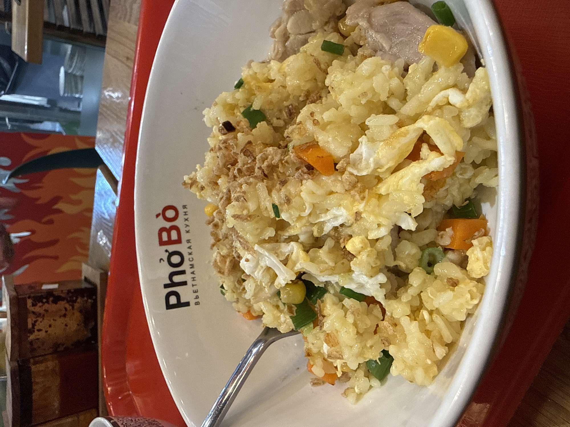 A close-up shot shows a bowl of fried rice, a dish often found in Vietnamese cuisine. The rice is mixed with various ingredients, including pieces of scrambled egg, corn kernels, green onions, and what appear to be fried shallots sprinkled on top. Some pieces of chicken are visible on the right side of the bowl. The bowl is white with the logo "Pho Bo" and "ВЬЕТНАМСКАЯ КУХНЯ" (Vietnamese Cuisine) printed in black and red on the rim. A silver fork is partially visible on the left, resting on the rim of the bowl. The bowl sits on a red tray, which is placed on a wooden table. In the background, out of focus, are elements of a restaurant setting, including condiment containers and a wall decoration with a flame motif. The overall lighting is warm and inviting.
The image depicts a food item in a restaurant. However, there is no visual information in the image that can be used to determine its location in Rostov-on-Don, Russia, or any other specific geographical location.