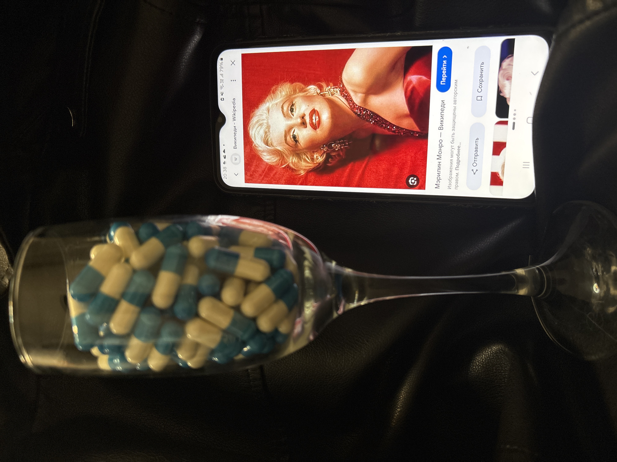 The image displays a clear glass champagne flute filled with numerous two-toned capsules, approximately uniform in size, featuring light blue and cream-colored halves. To the right of the flute, a black smartphone is positioned vertically, its screen displaying a Wikipedia page for "Мэрилин Монро" (Marilyn Monroe). The prominent image on the screen is a color portrait of Marilyn Monroe with blonde hair, red lipstick, and a red dress set against a red background. The phone's status bar shows the time as 20:38, Wi-Fi and LTE indicators, and a 79% battery charge. The objects rest on a dark, textured surface, likely black leather, exhibiting creases and reflections. The specific location within the city of Izhevsk, Russia, cannot be determined from the visual information presented in the image.