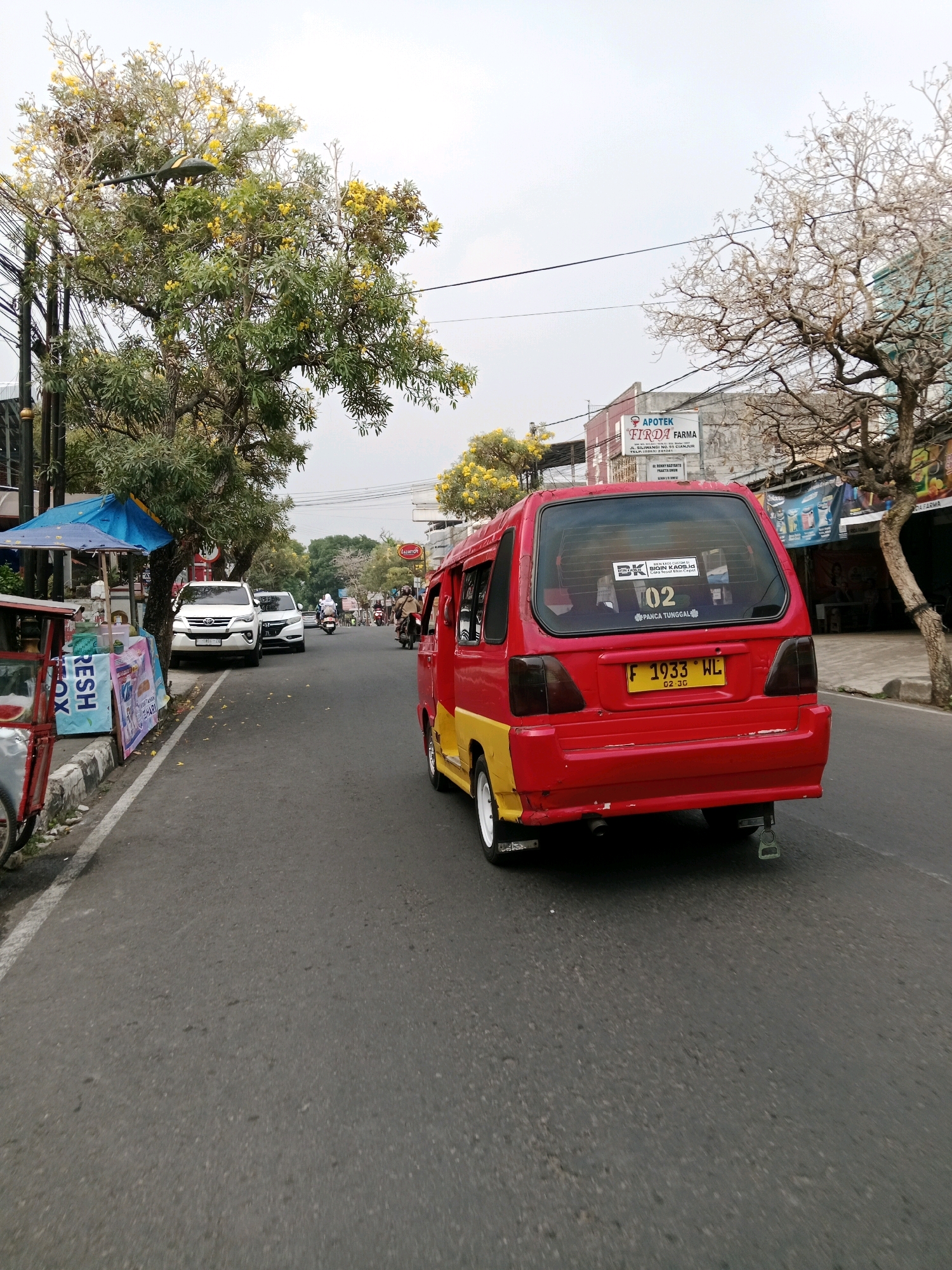This image captures a street scene in Cianjur, Indonesia, likely during daylight hours. The foreground is dominated by the rear of a red and yellow van, with a yellow license plate displaying "F 1933 WL". A sticker with the text "BK" and "02" is visible on the rear window, along with "PARCA TUNGGAL". The van is traveling on an asphalt road with a white line marking the edge.
To the left of the road, there is a sidewalk with a small blue umbrella over a vendor's cart, which displays "FRESH" prominently on a banner. Behind the cart are two white SUVs parked on the side of the road, one with license plate "F 1680 VJ". Tall trees with yellow flowers are lining the street, obscuring some of the buildings in the background. One building features a sign that reads "APOTEK FIRDA FARMA". In the distance, other vehicles, including motorcycles and a white car, are visible, along with pedestrians. The sky is overcast.